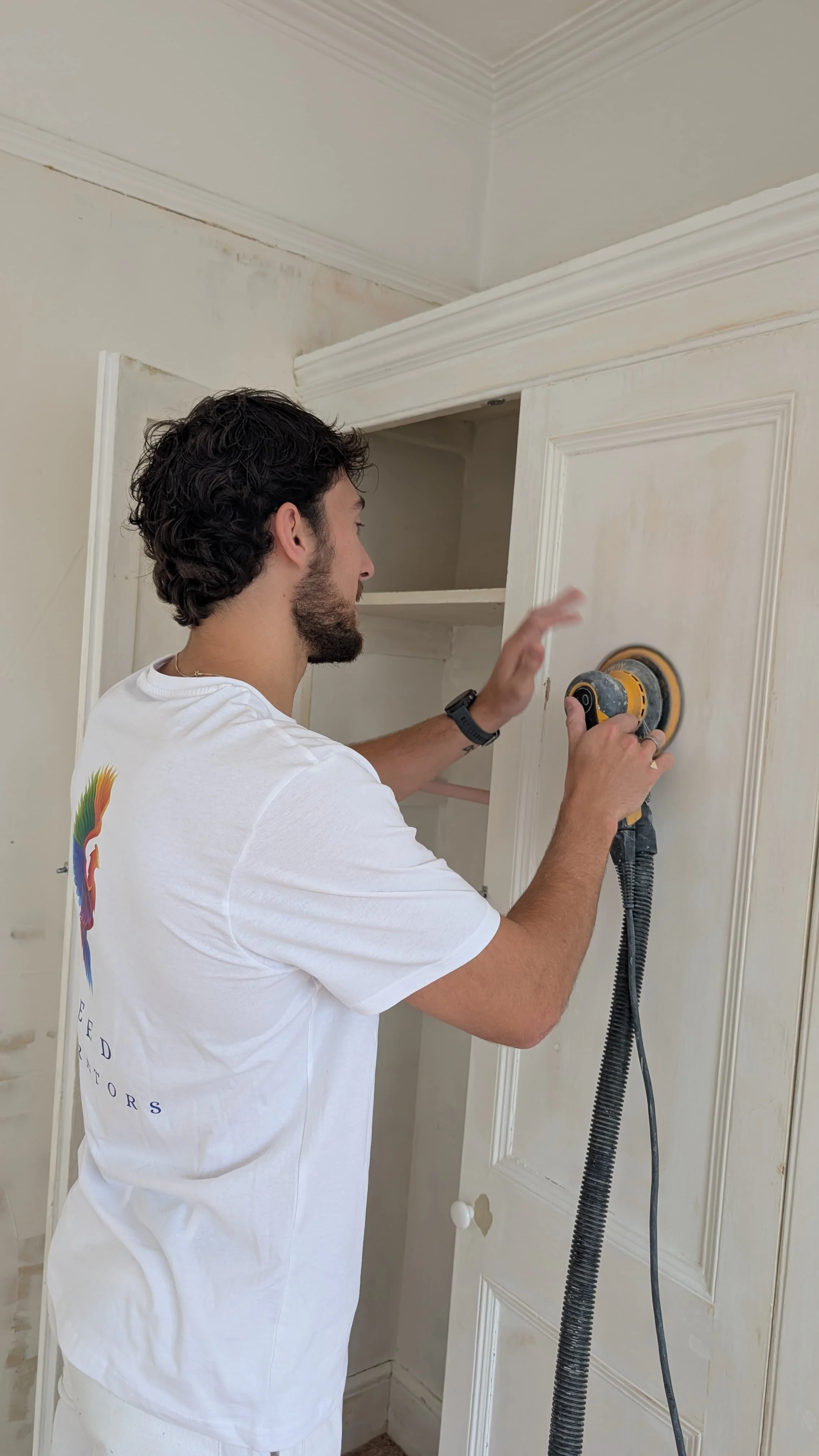 A man with dark curly hair and a beard wearing a white t-shirt sanding a door with a power sander in a room.