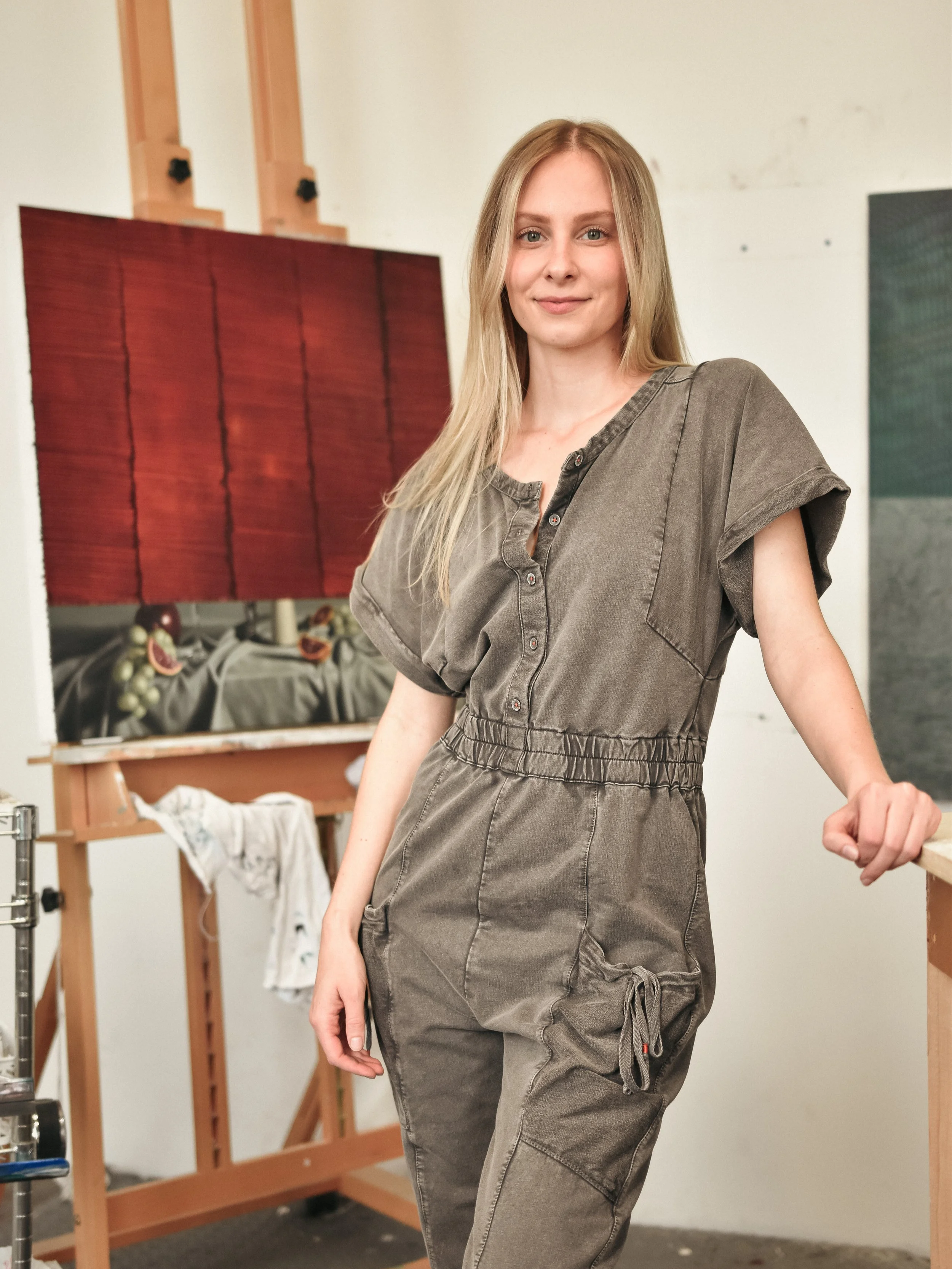 una giovane donna con capelli biondi e occhi chiari indossa un tutone grigio mentre si trova in un'atelier di pittura con tele e materiali artistici sullo sfondo