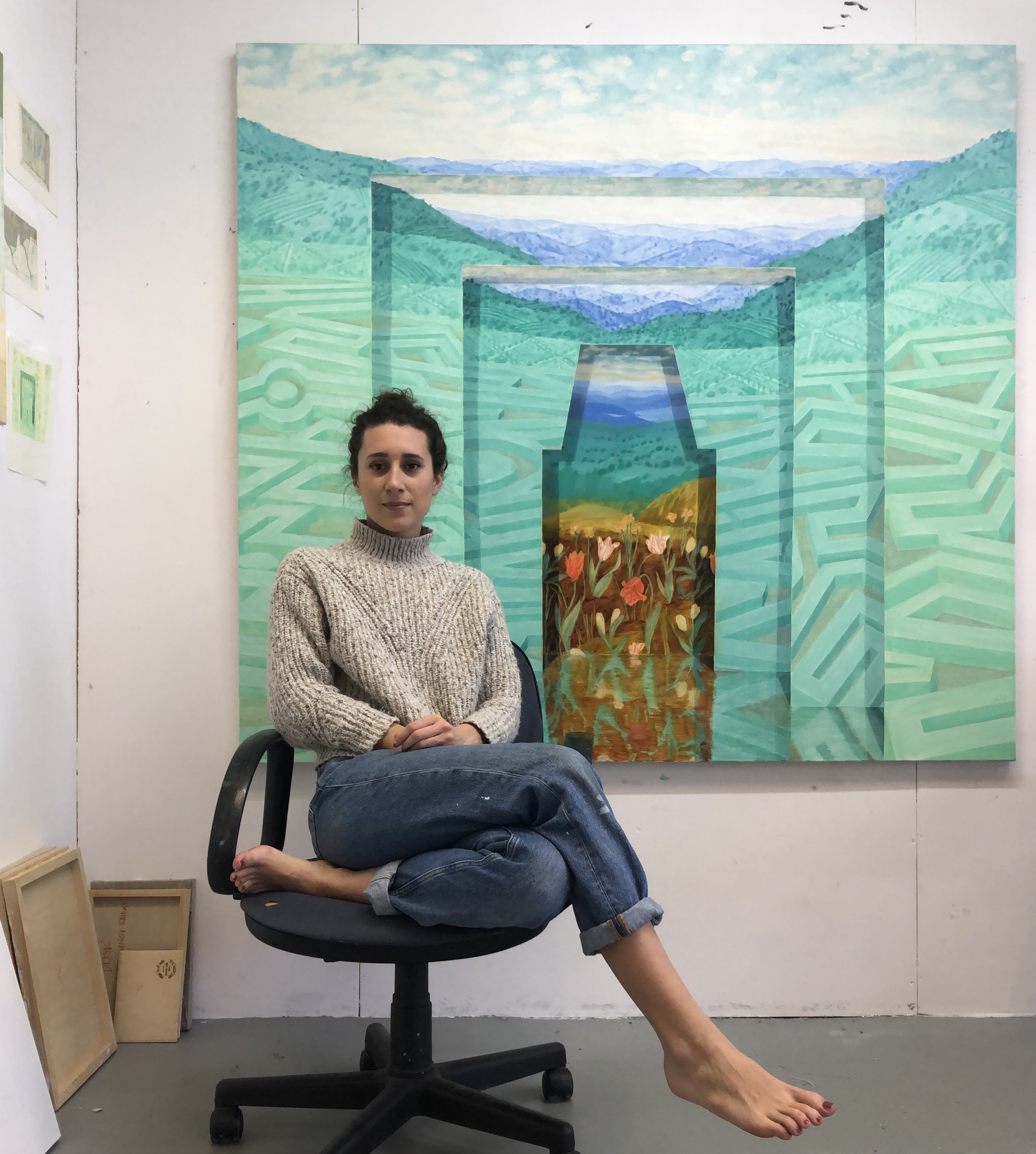 Una donna seduta su una sedia con un quadro alle spalle di un paesaggio colorato e stilizzato, e alcuni telai di legno accatastati a sinistra.