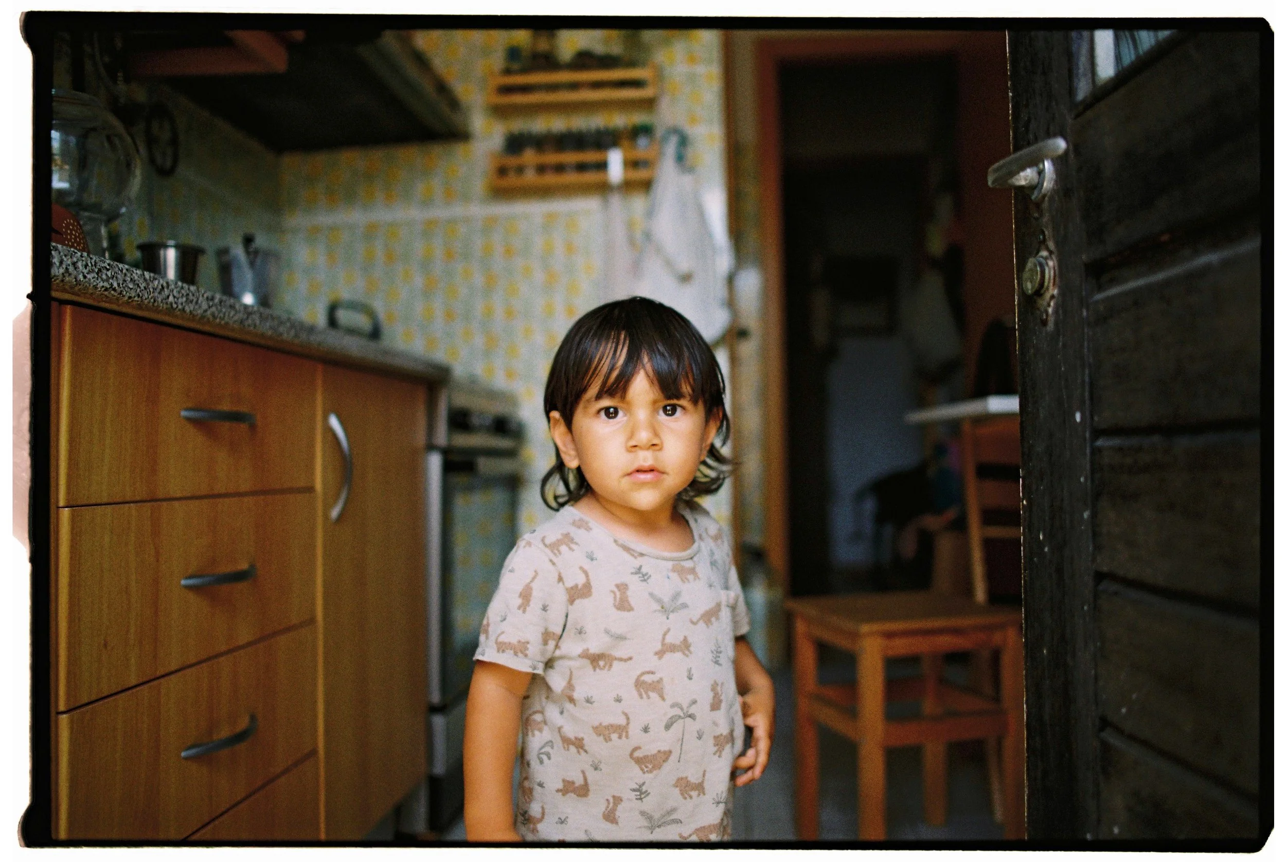 A young girl with dark hair, wearing a beige shirt with animal prints, stands in a kitchen with wooden cabinets and a tiled backsplash, looking at the camera.