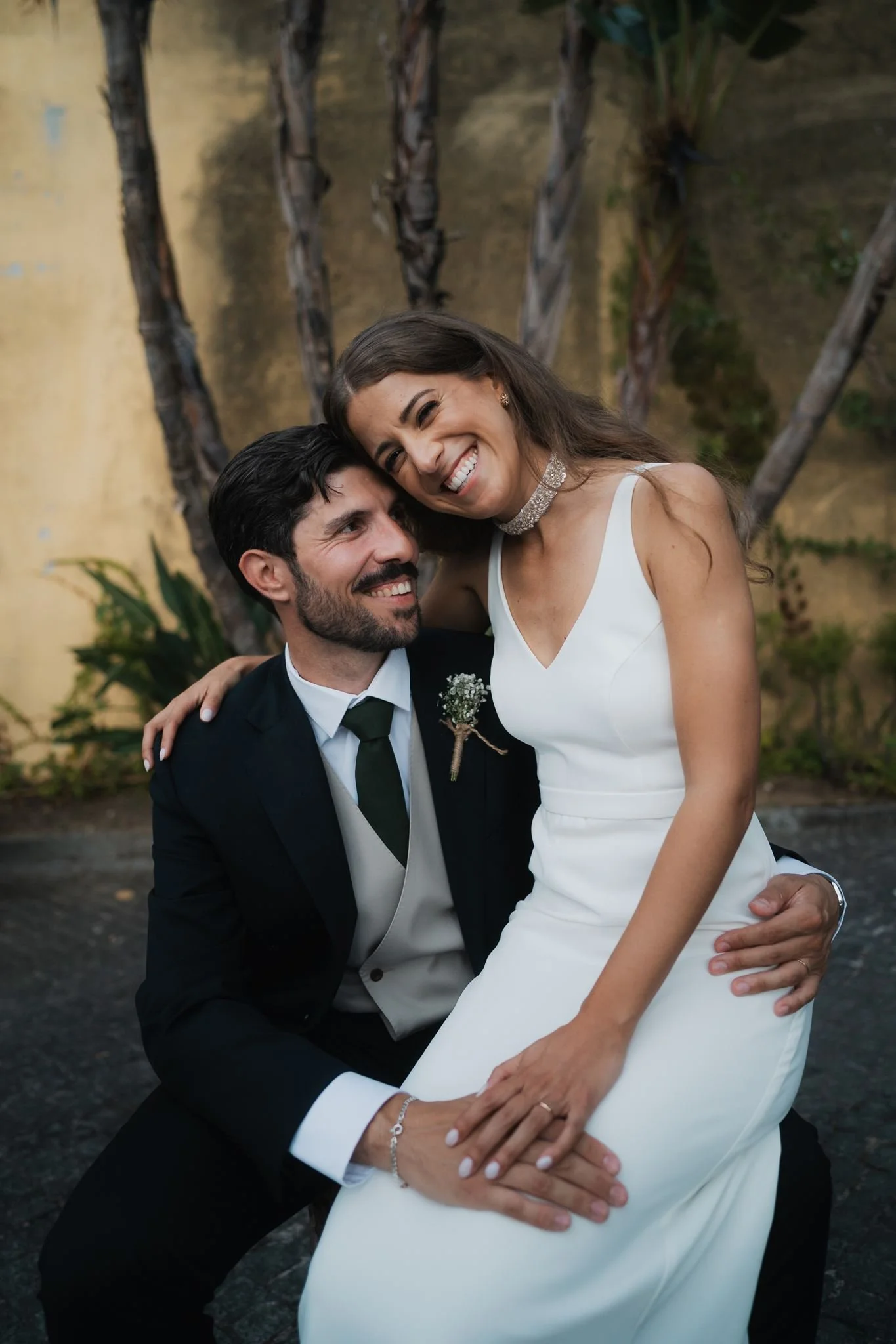 A couple dressed in wedding attire, smiling and embracing outdoors with trees and foliage in the background.