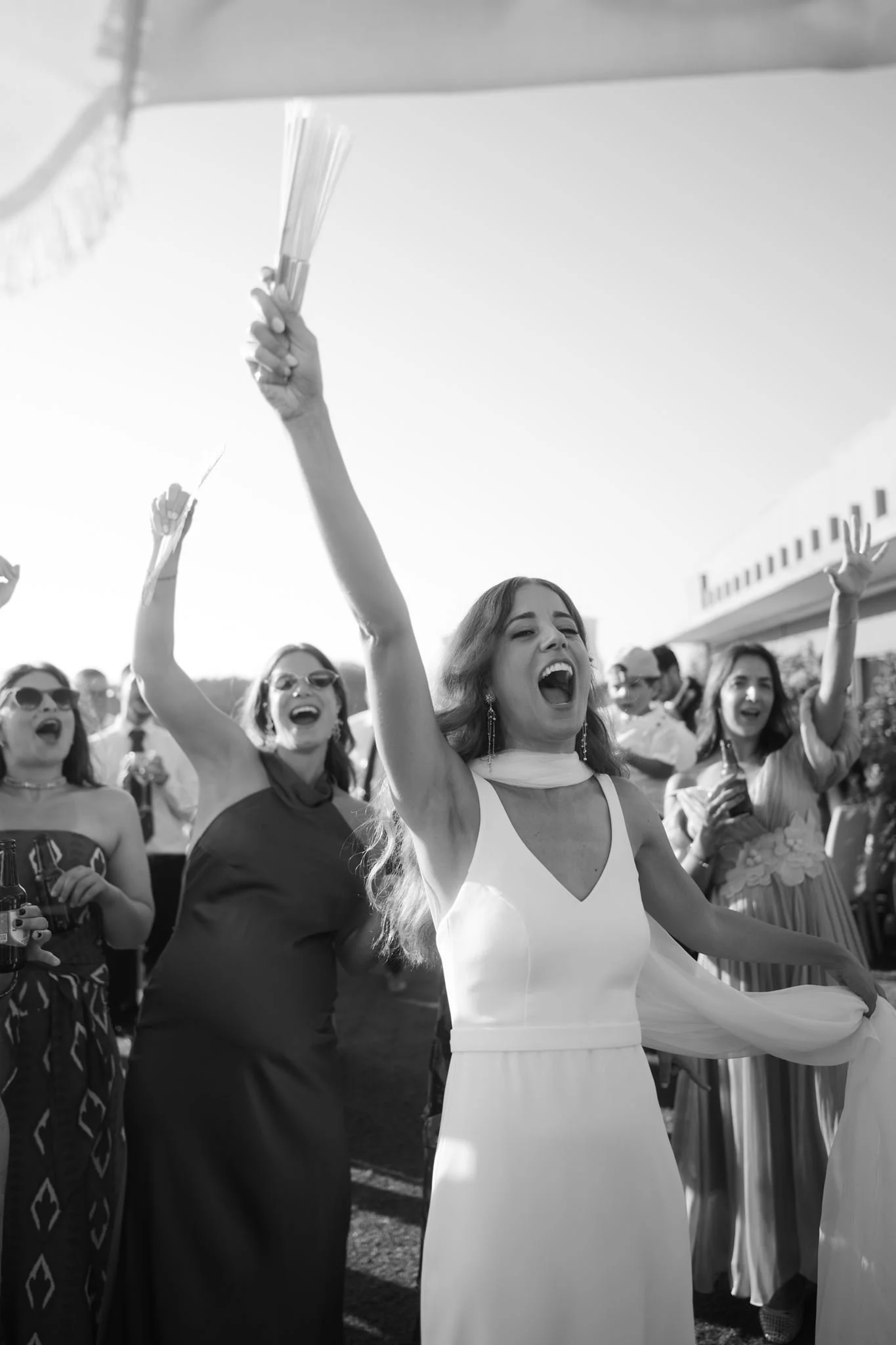People celebrating outdoors, smiling, and raising their hands, with one woman in the foreground wearing a white dress.