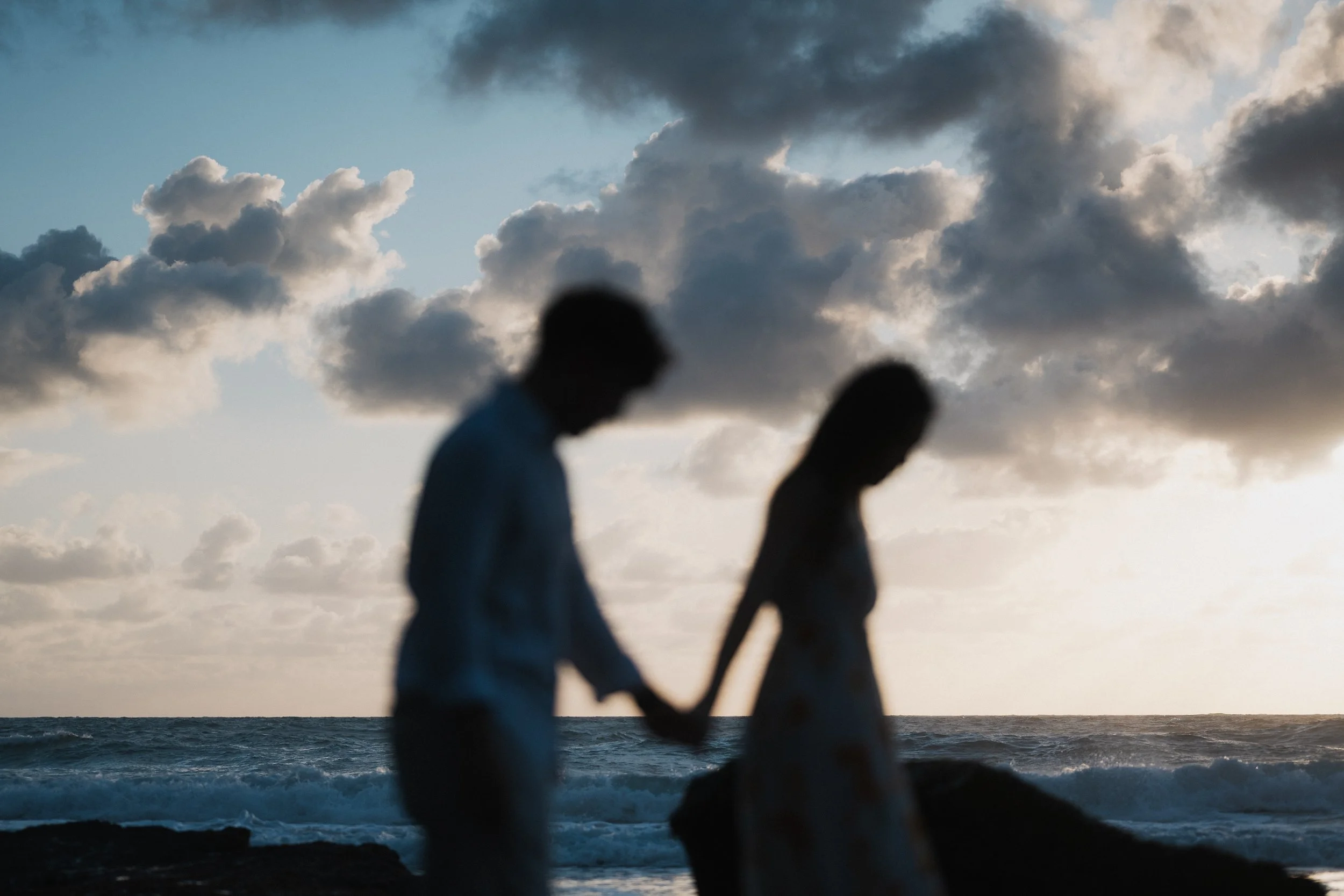 Silhouettes of two people walking hand in hand on a beach with cloudy sky and ocean in the background.