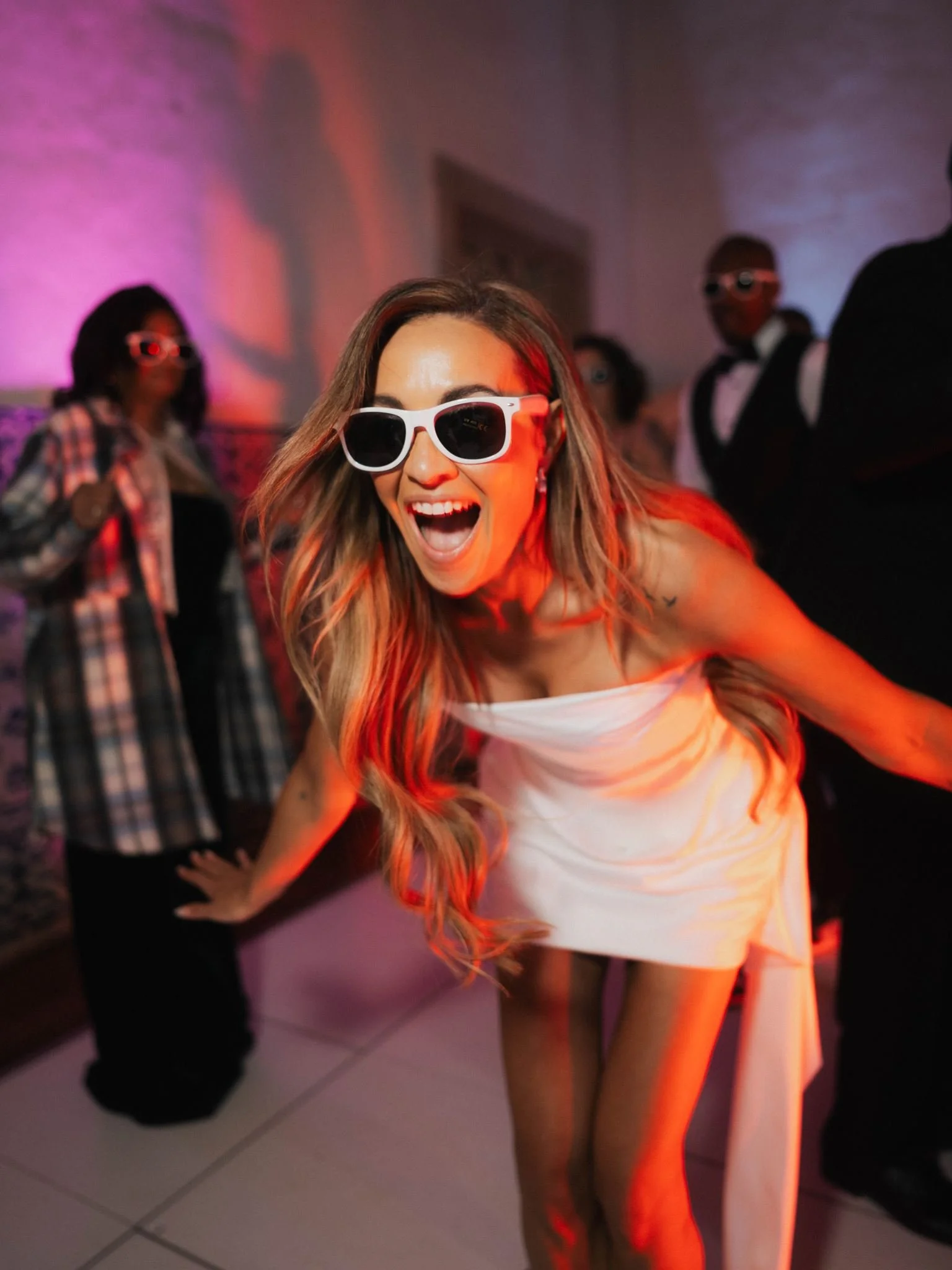 A woman with long hair wearing white sunglasses and a strapless dress, smiling widely and dancing at a party with colorful lighting. In the background, other people are visible, including a woman in a plaid shirt and sunglasses, and a man in a tuxedo.