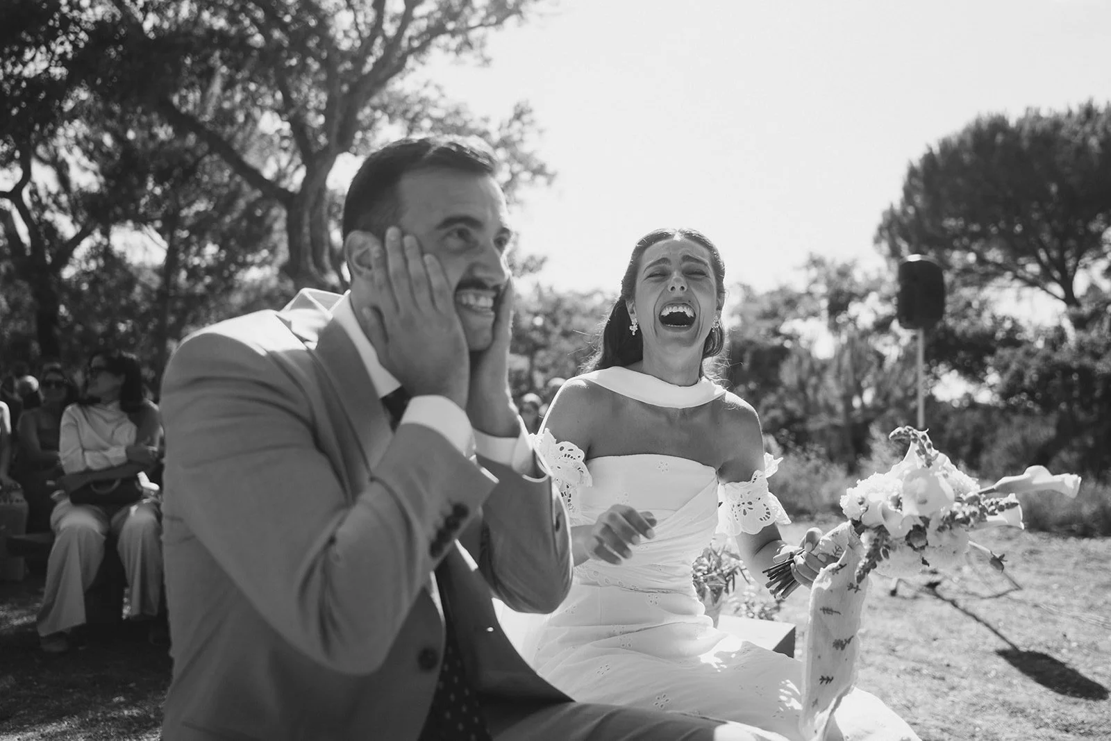 A black-and-white photo of a bride and groom at their wedding, both laughing joyfully outdoors. The bride holds a bouquet of flowers, and the groom has his hands on his cheeks, smiling widely, during their wedding ceremony at Quinta dos Anjos, captur