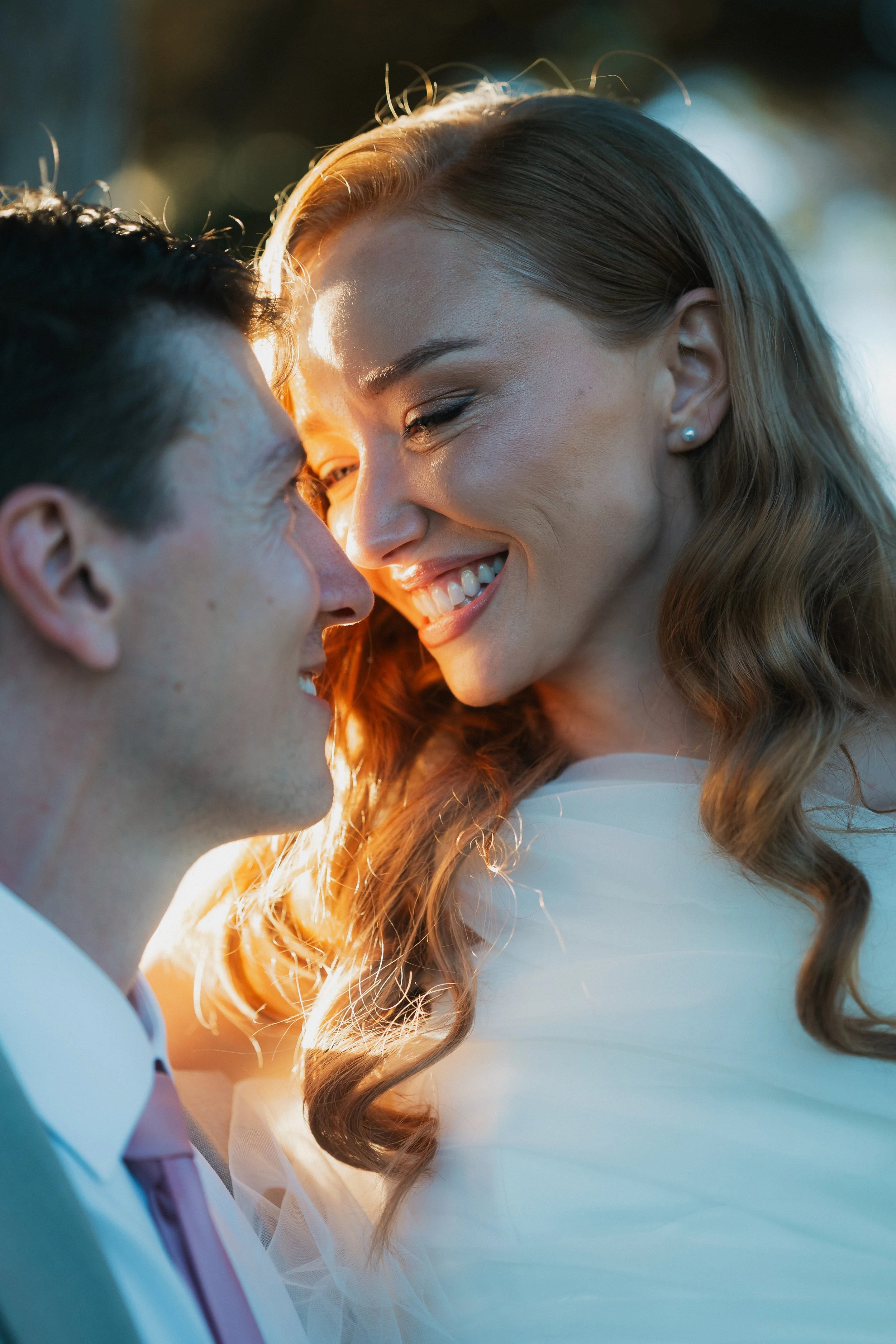 Close-up of a smiling couple about to kiss, with the woman having long, curly hair and the man with short, dark hair, in a warm, golden light.