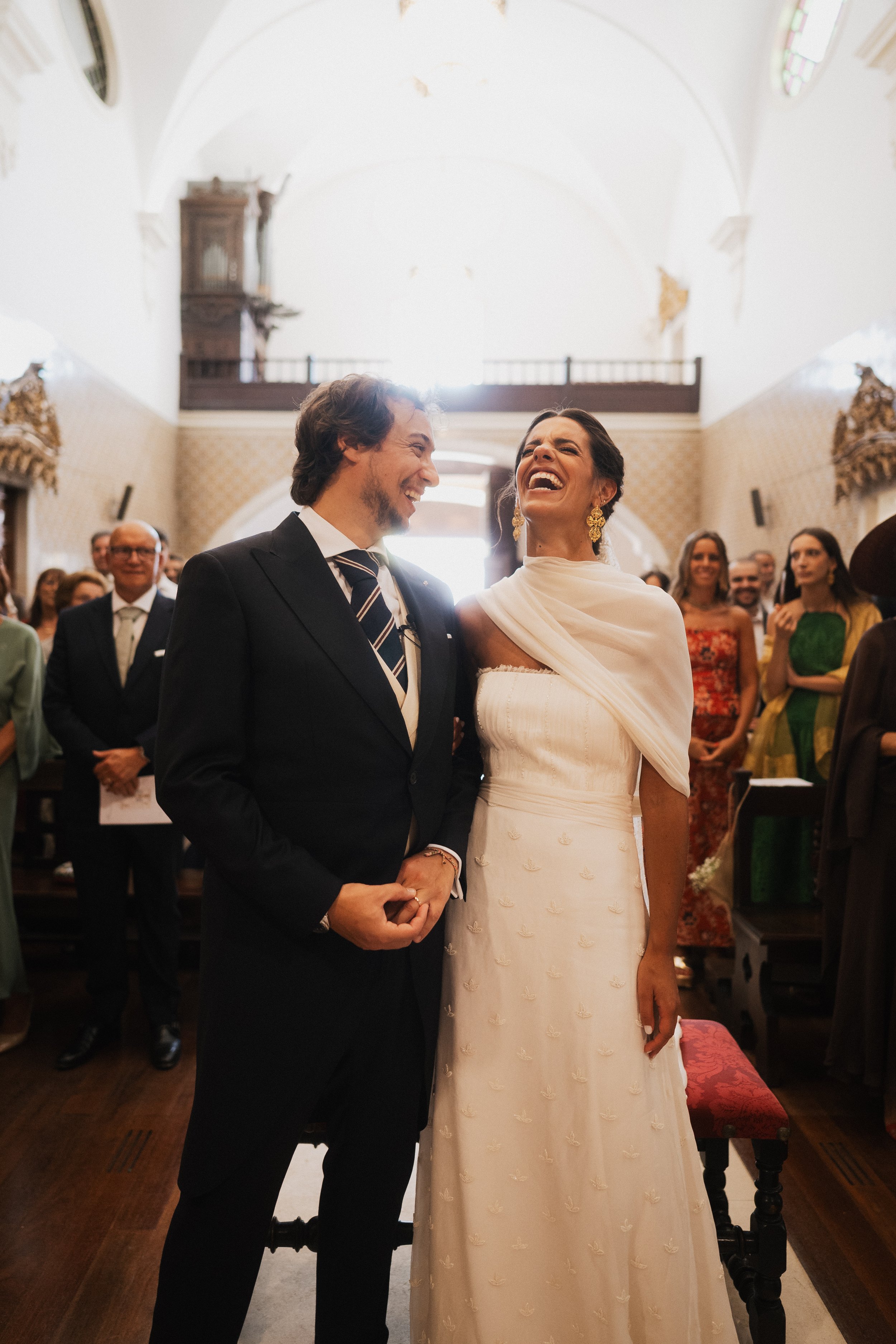 A wedding ceremony inside a church with a bride and groom holding hands and smiling at each other, surrounded by guests.