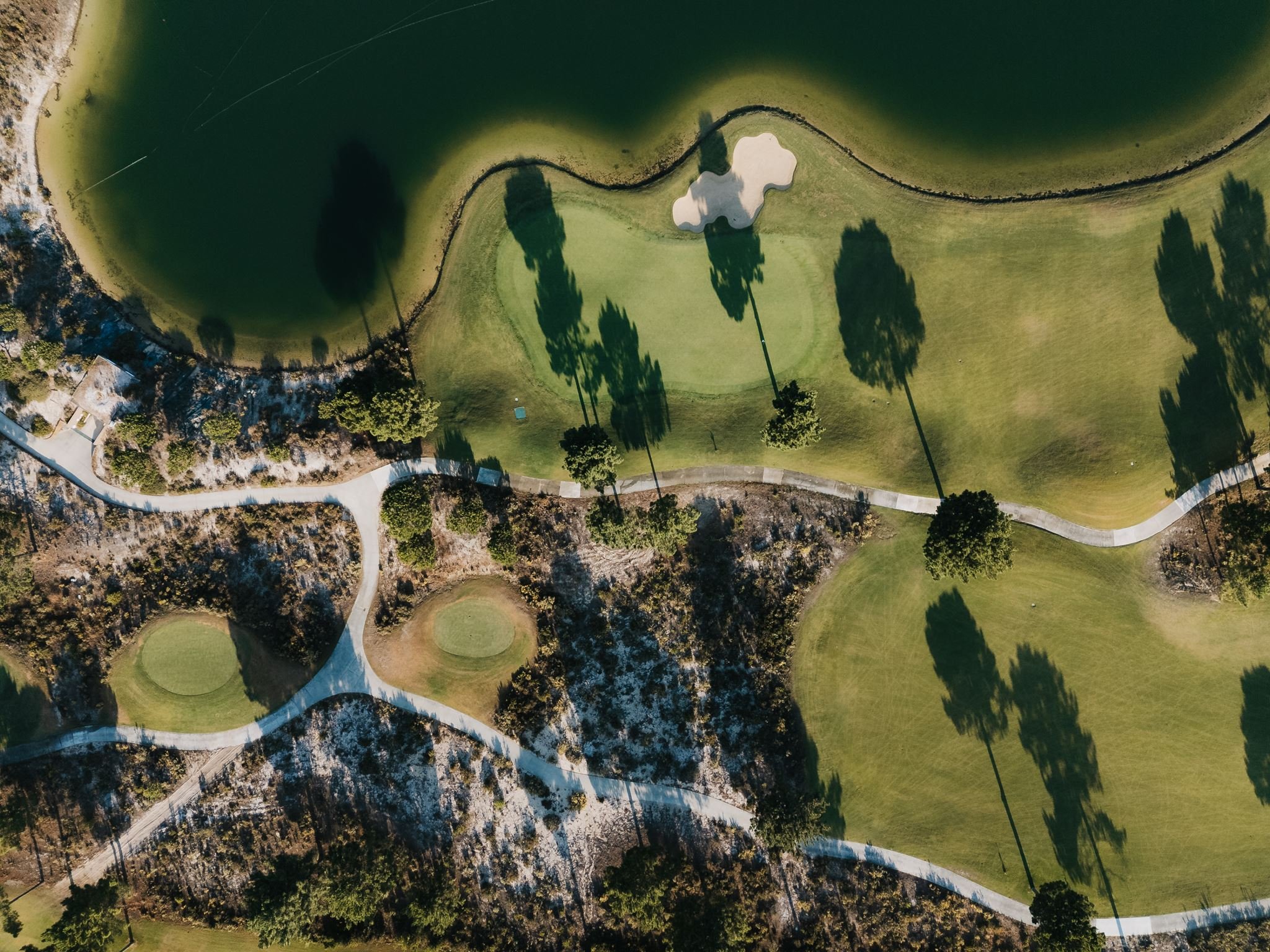 An aerial view of a golf course featuring a putting green, a sand trap, water hazards, trees, pathways, and surrounding natural landscape.