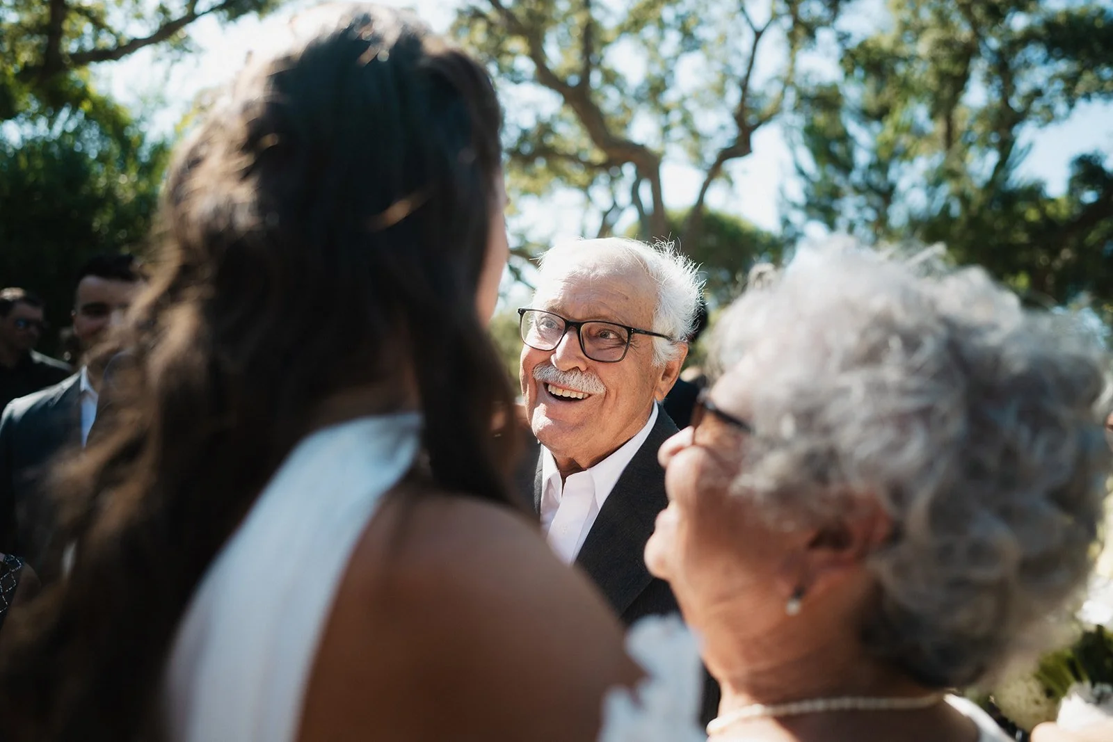 Three women and two men are smiling and talking outdoors, with trees in the background. The man with glasses and a white shirt is in the center, smiling at the women.