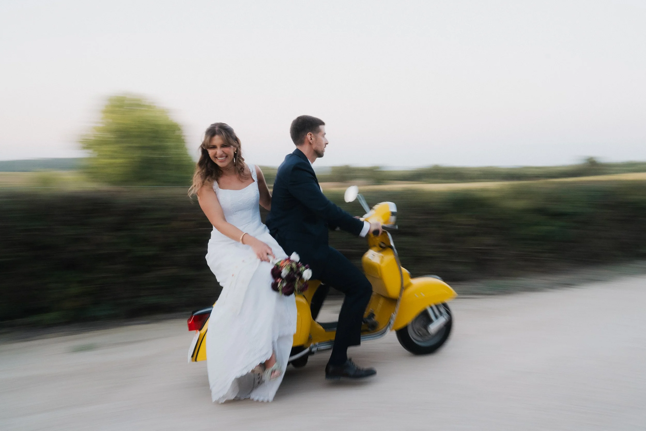 A couple dressed in wedding attire riding a yellow scooter together down a rural road, with the bride holding a bouquet of flowers and both smiling and enjoying the ride.