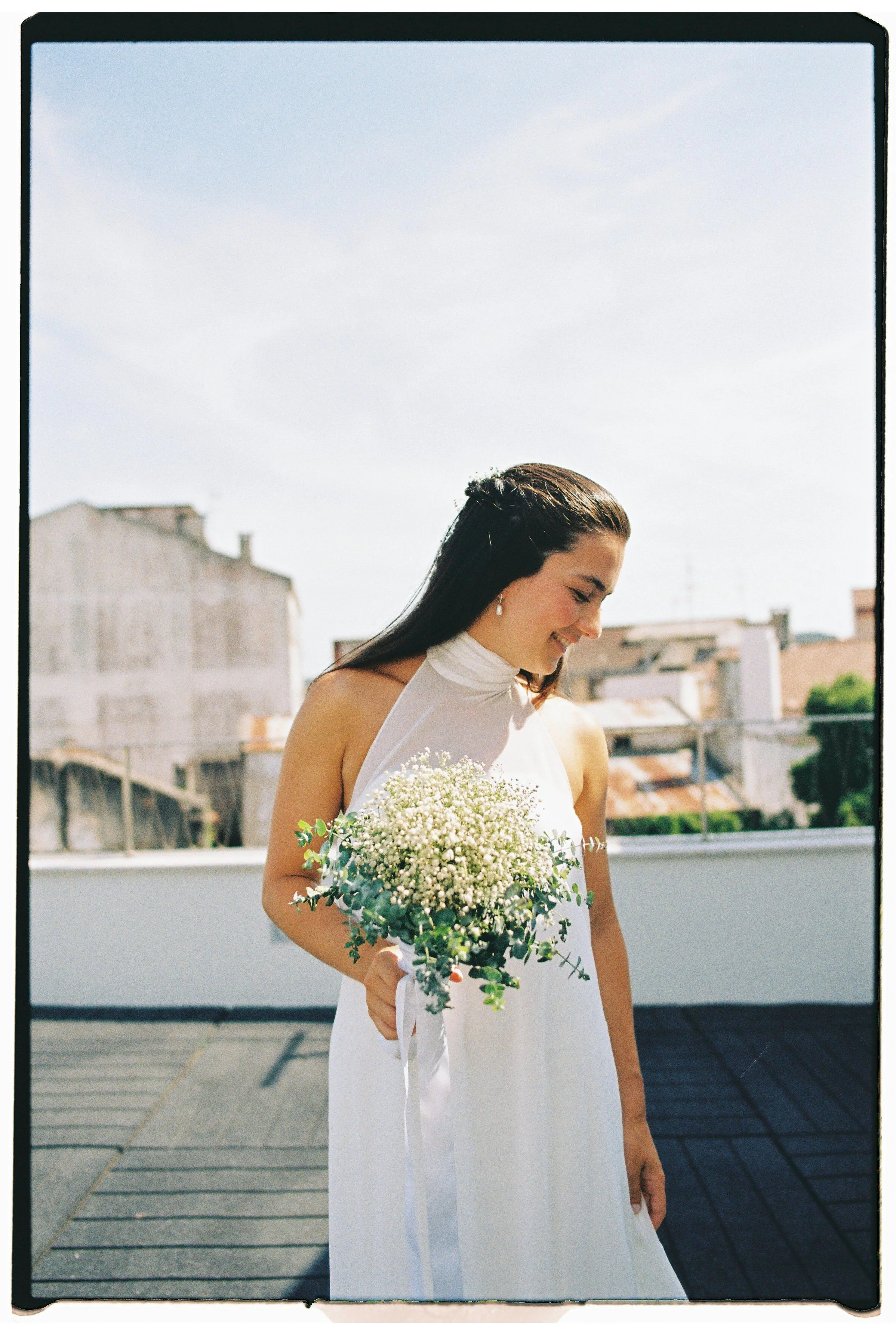 A smiling woman in a white dress holding a bouquet of white flowers outdoors on a rooftop with buildings in the background.