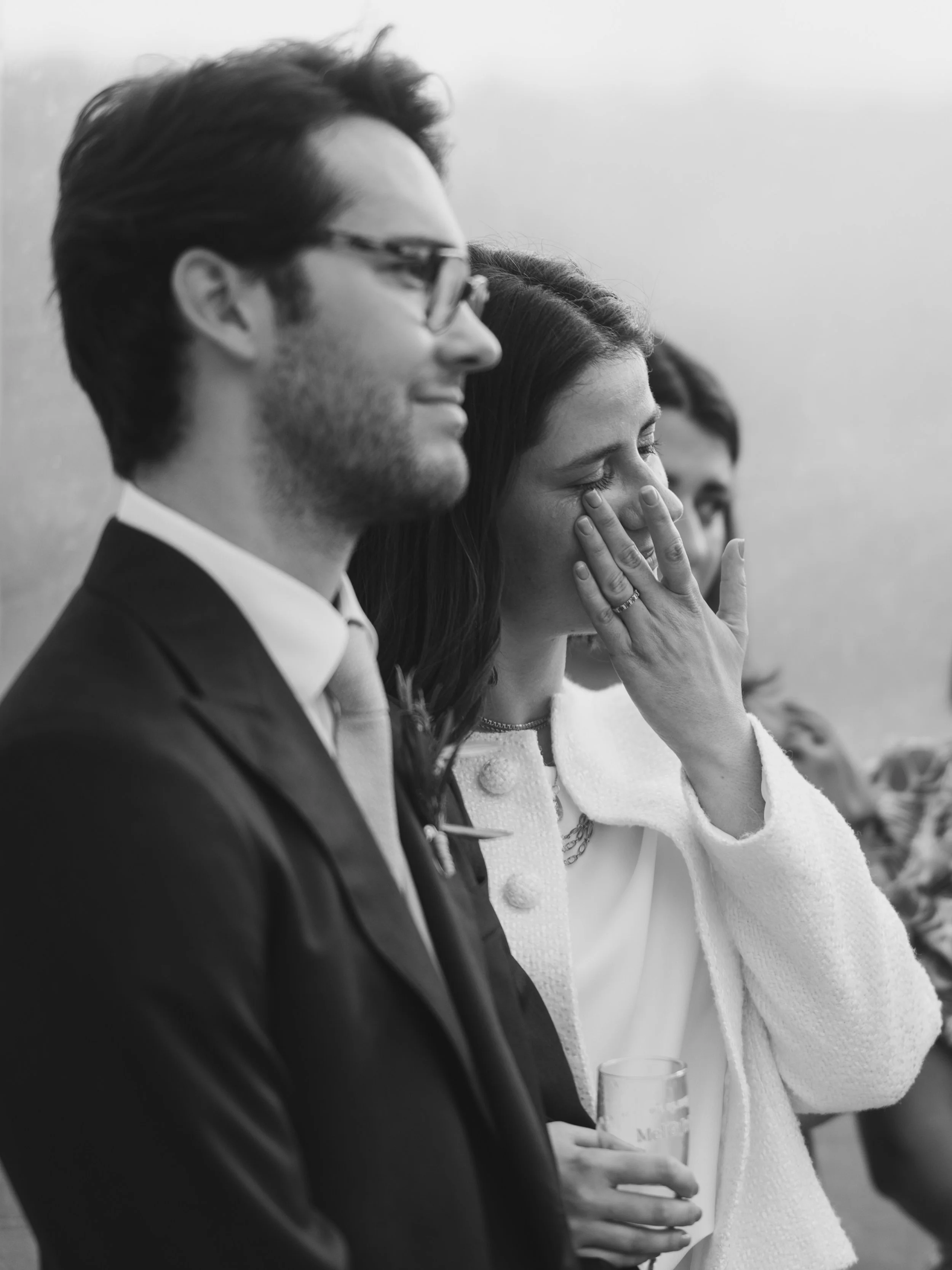 A black and white photo of three people standing side by side, with the woman in the middle covering her face with her hand and closing her eyes, while the men on either side are looking forward, all with relaxed expressions.