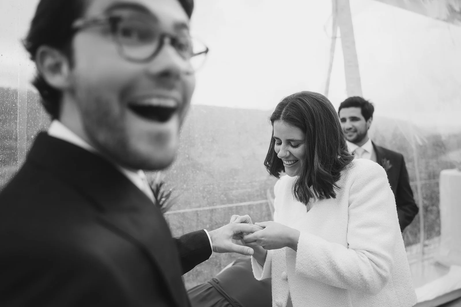A woman appears to be putting a ring on a man's finger during a wedding ceremony, with a man in a suit smiling in the background.