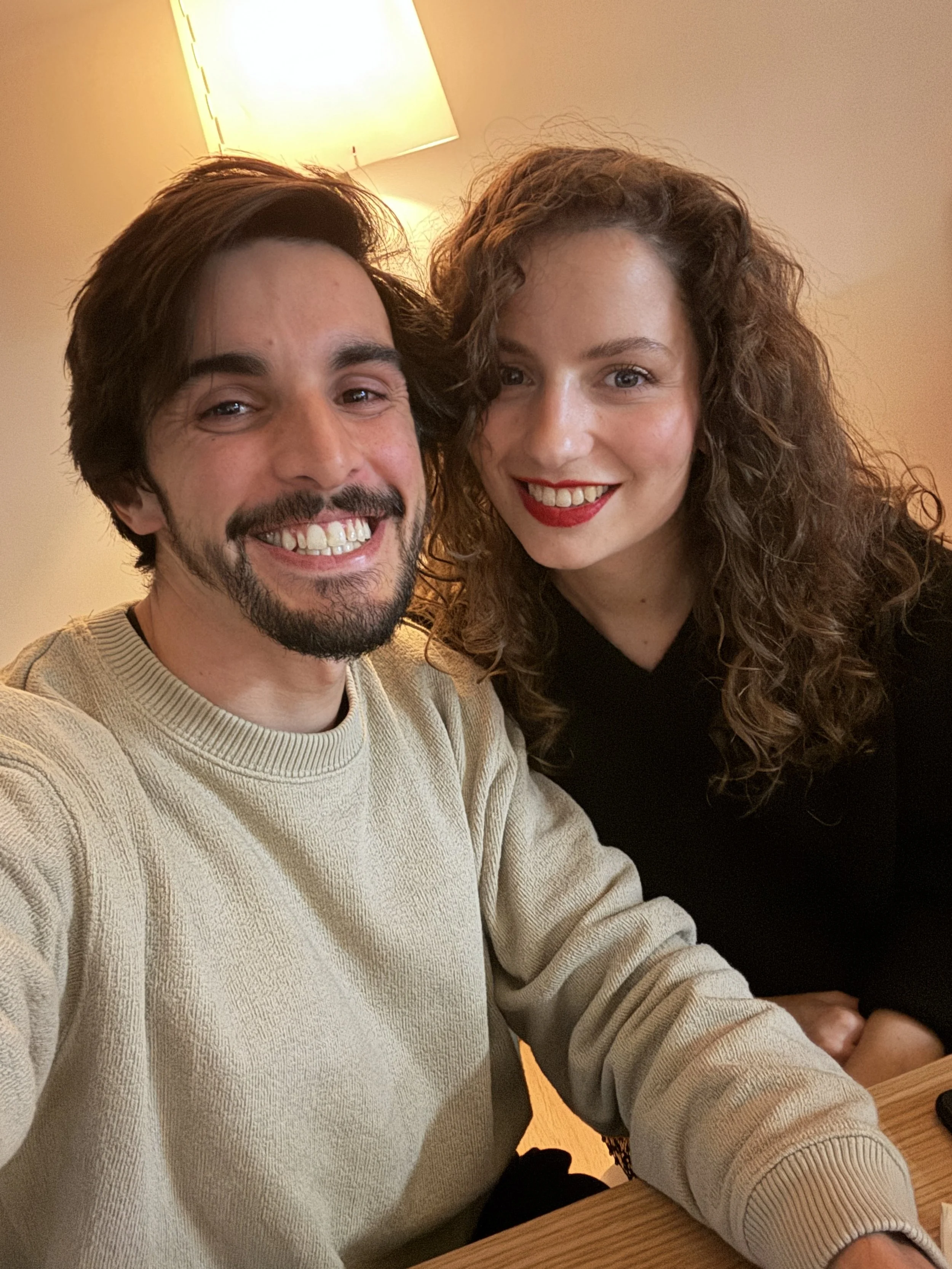 A man and a woman smiling in a warmly lit room, taking a selfie together.
