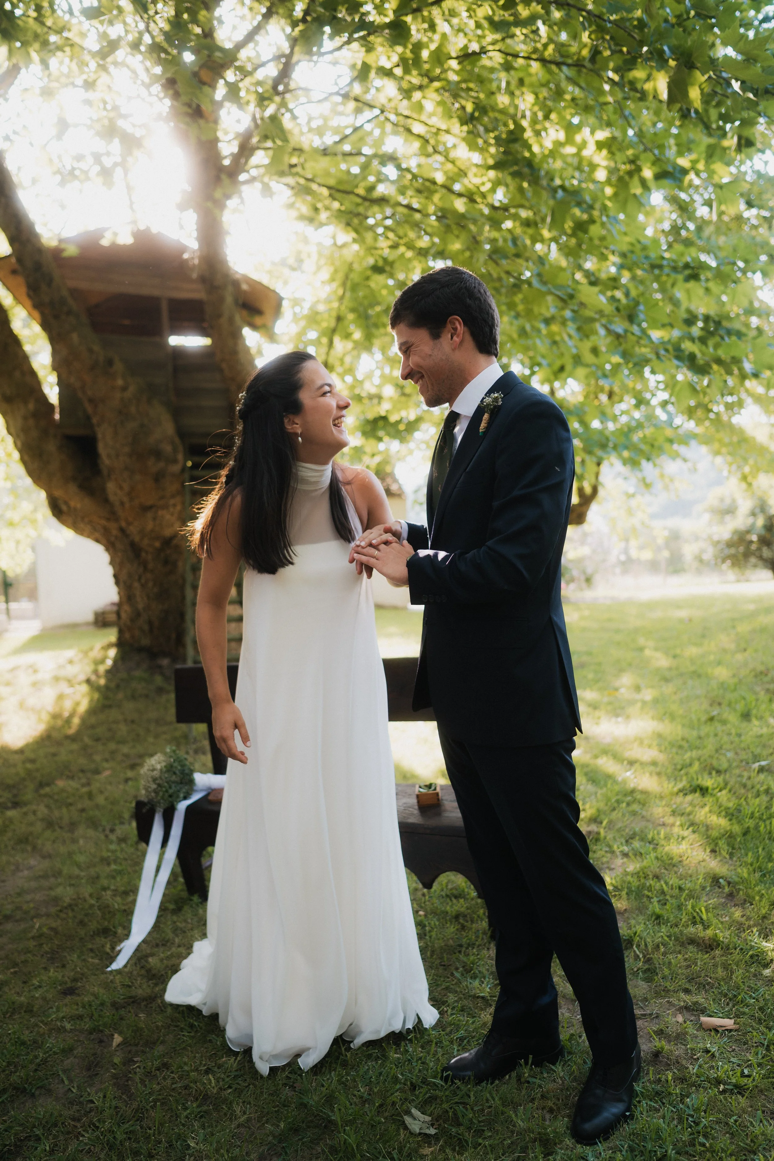 A bride and groom holding hands and smiling during their outdoor wedding ceremony under green leafy trees.