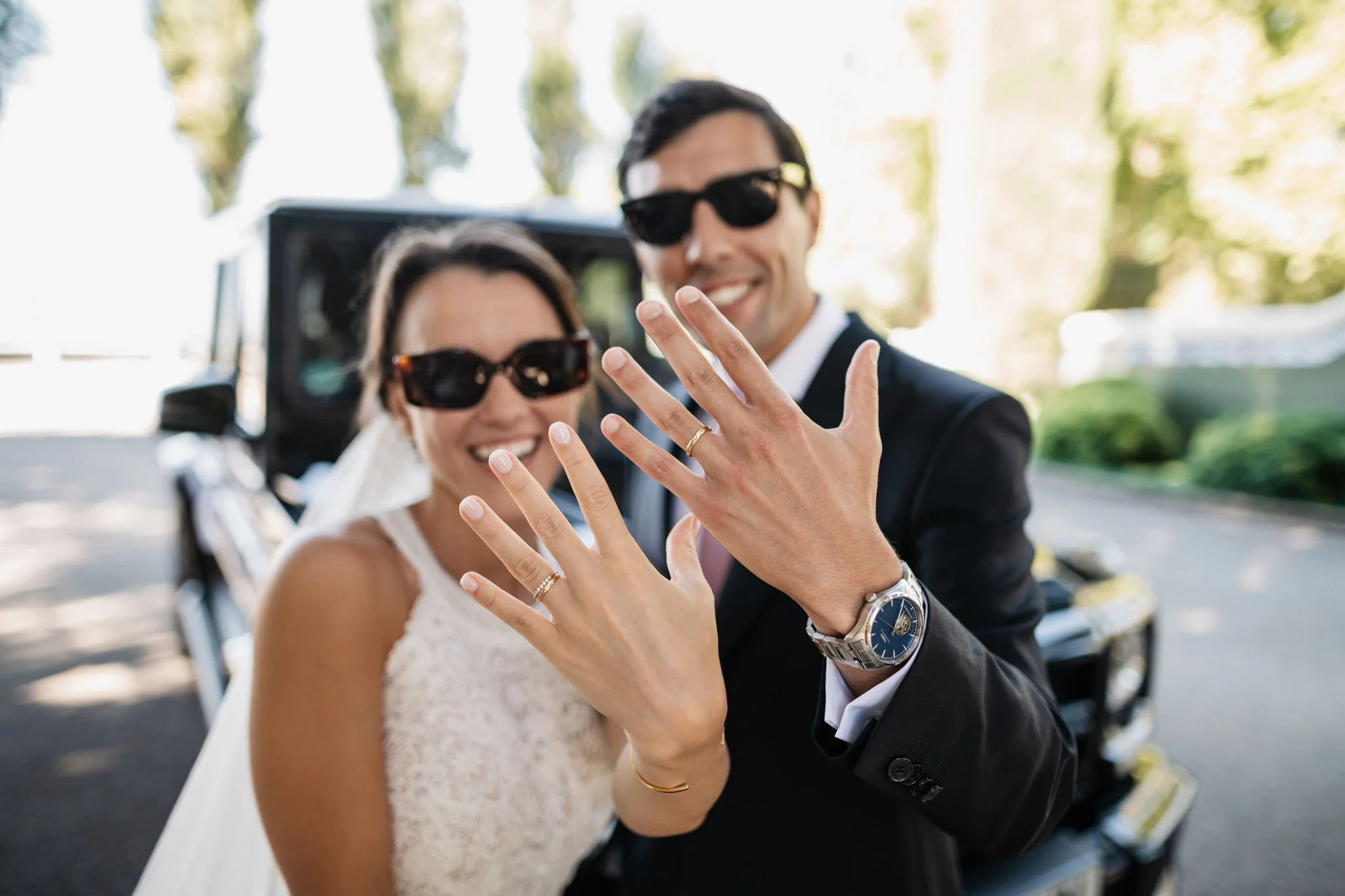 A newlywed couple showing their wedding rings, wearing sunglasses and smiling, in front of a parked car.