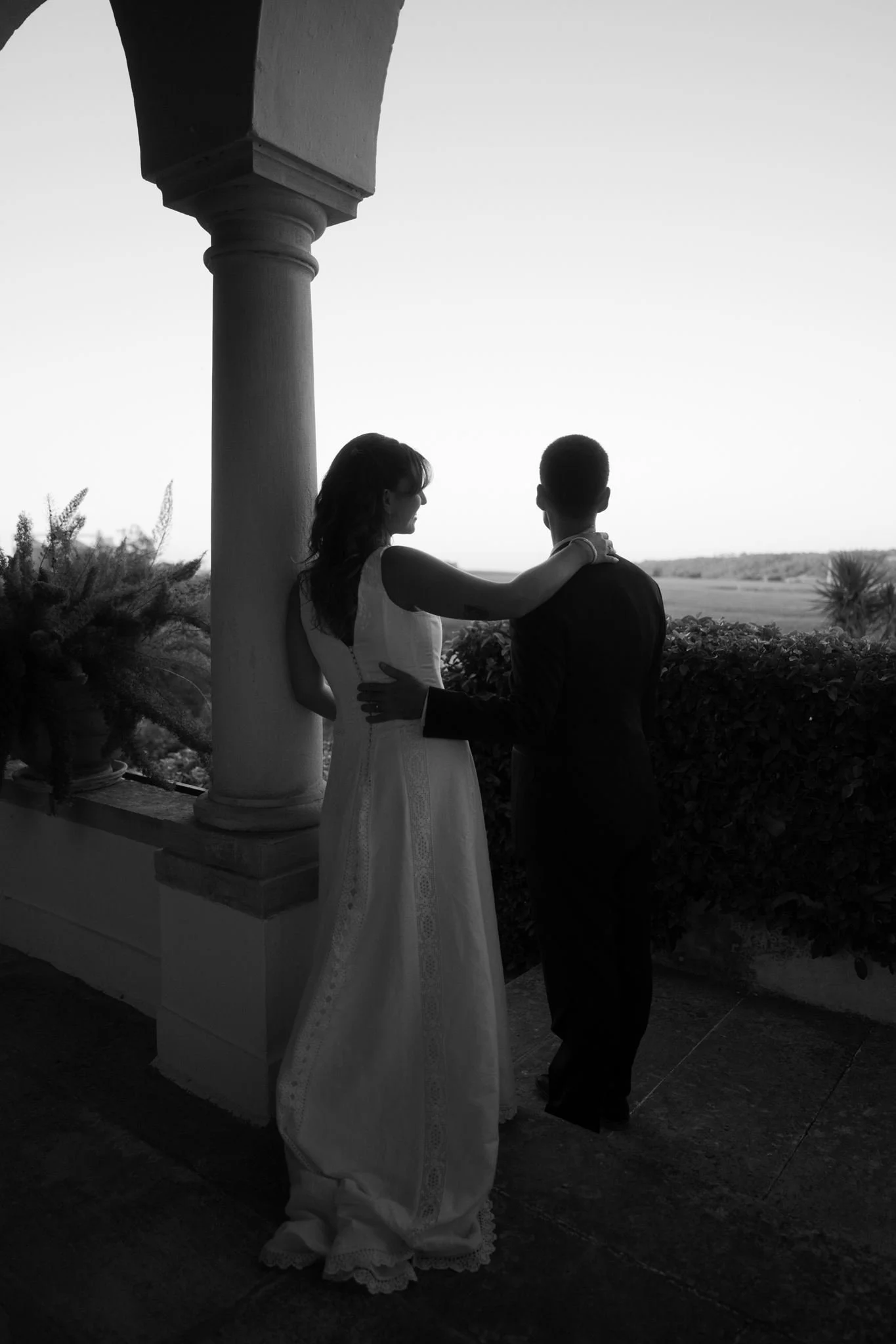 A black and white photo of a couple standing outdoors near a column, facing each other, with the woman resting her arm on the man's shoulder and holding his hand, overlooking a landscape with bushes and trees in the distance, during sunset or sunrise.