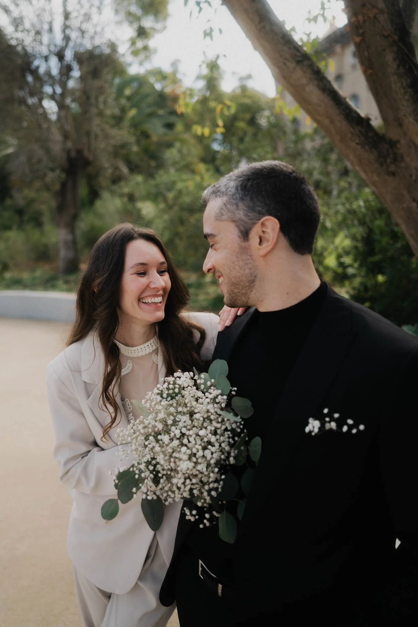 A couple smiling at each other outdoors, holding a bouquet of flowers.