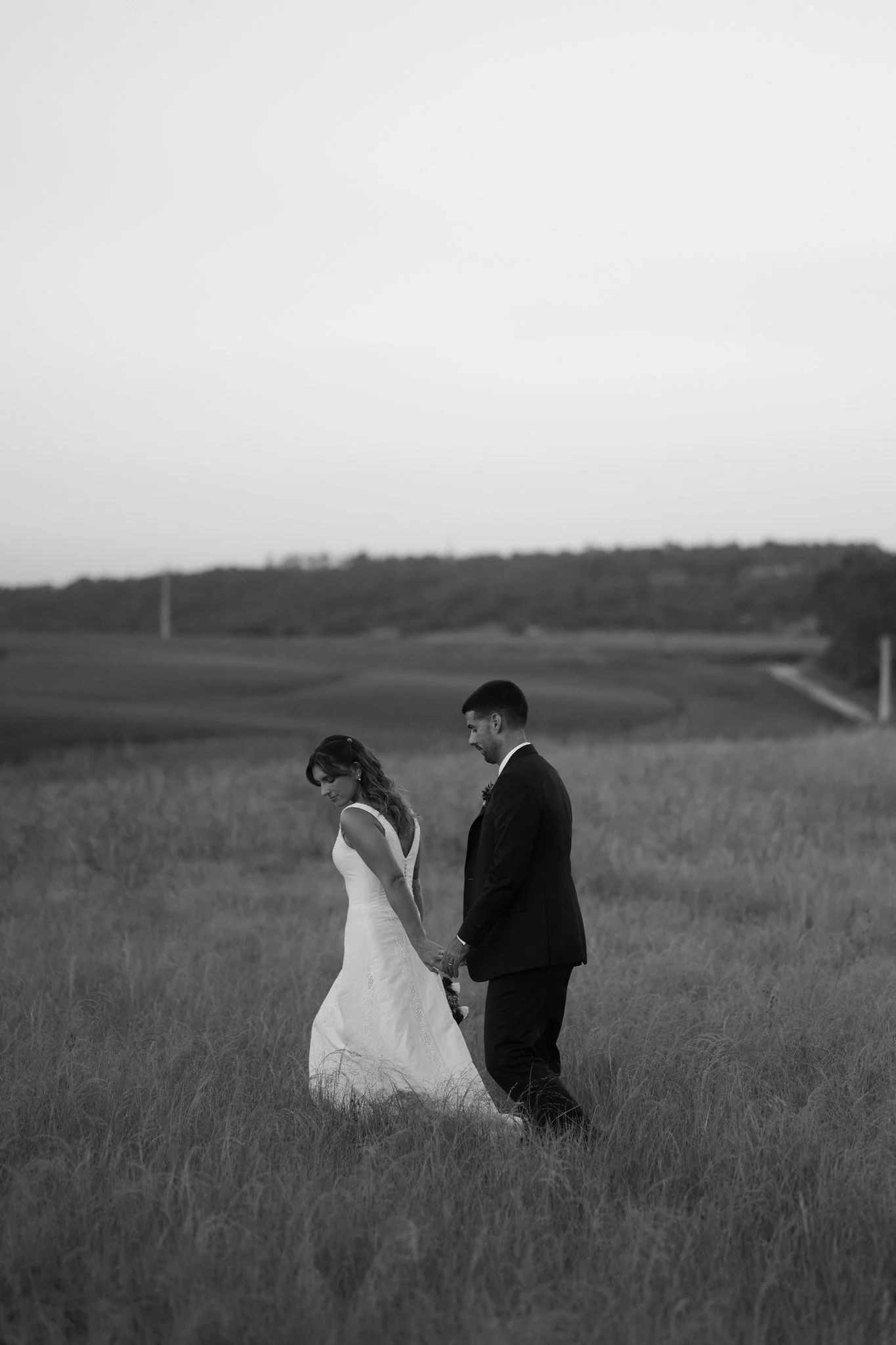 A black and white photo of a bride and groom holding hands and walking away in a grassy field with a hill in the background, beside a tree.