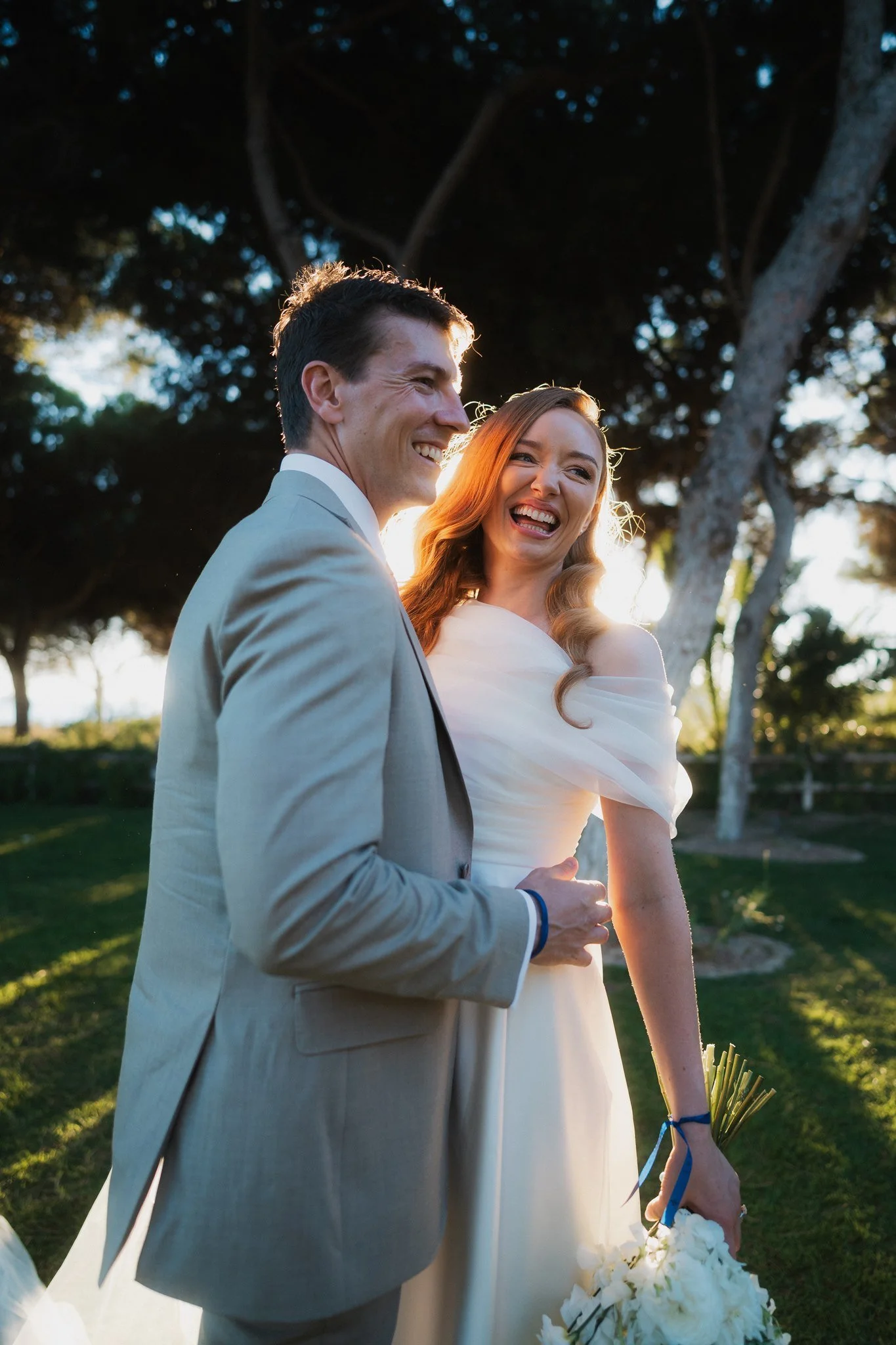 A newlywed couple smiling and laughing outdoors during sunset, standing close together, with the bride holding a bouquet of white flowers and the groom in a light gray suit.
