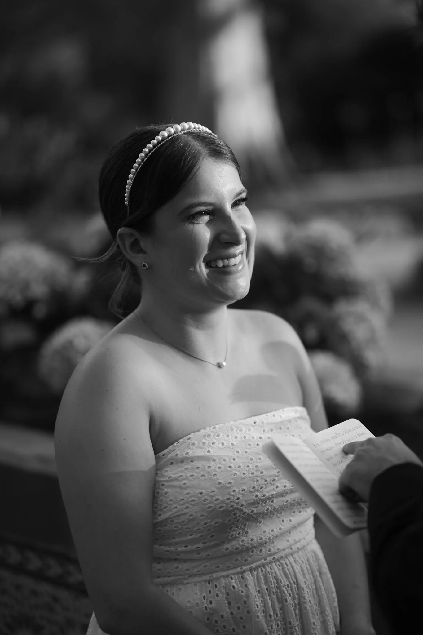 A smiling woman dressed in a strapless eyelet dress with a pearl headband and jewelry holds a handwritten note, with a person taking her photo in the background.