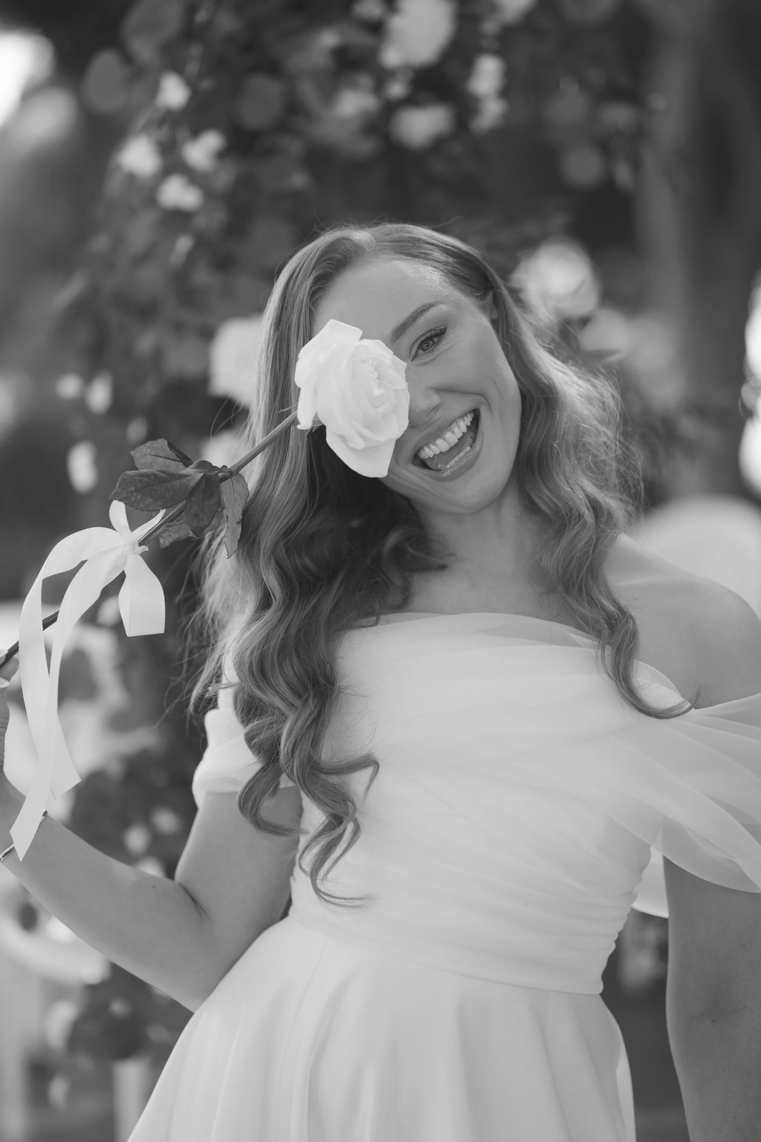 A woman with long wavy hair smiling and holding a white flower near her face, standing outdoors with a blurred background of trees and sunlight.