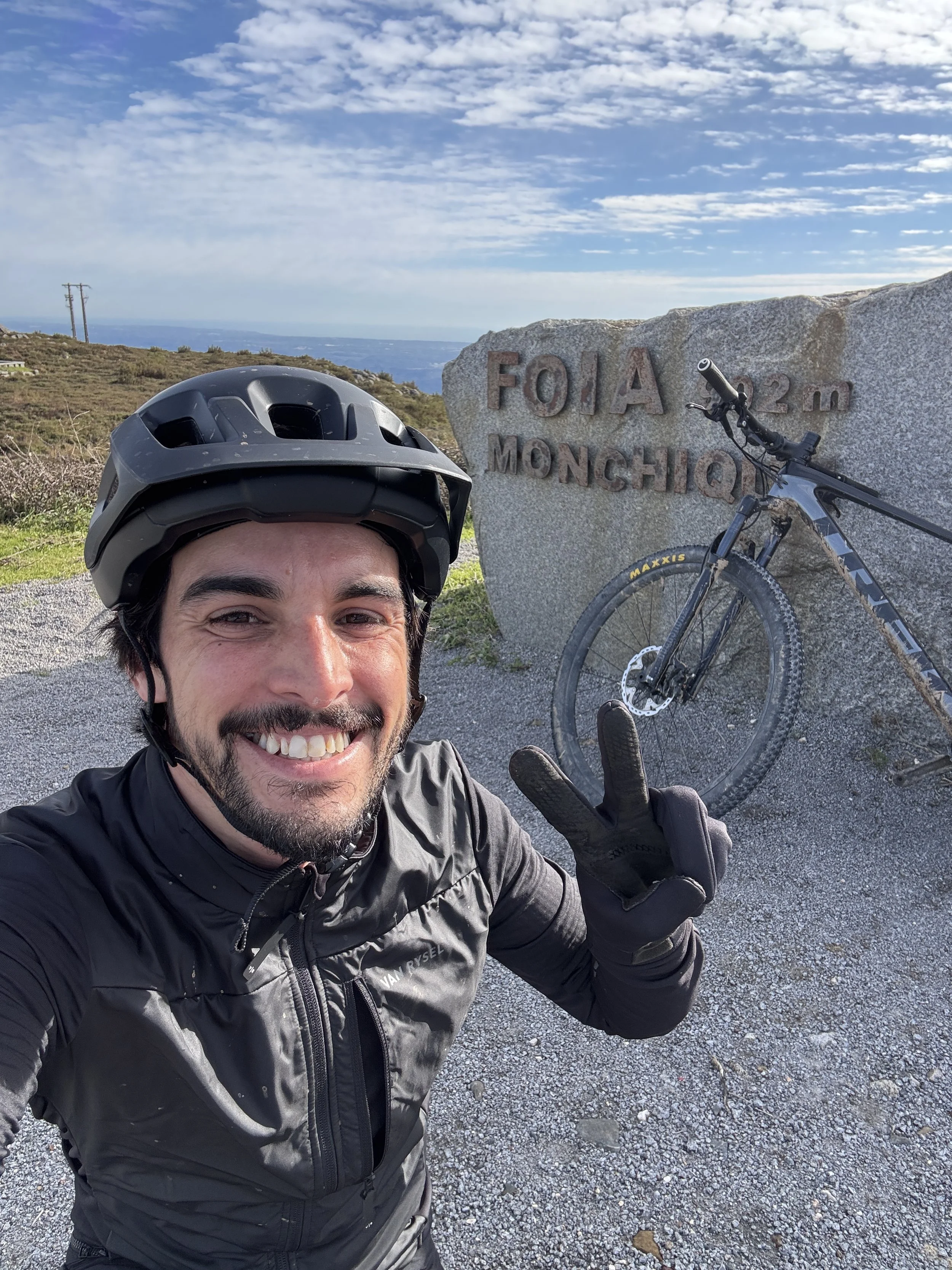 A cyclist taking a selfie at Foia Monchique, a mountain peak 923 meters high in Portugal. The cyclist is smiling, wearing a helmet and a black jacket, and making a peace sign. A mountain bike is leaning against a large stone marker with the inscripti