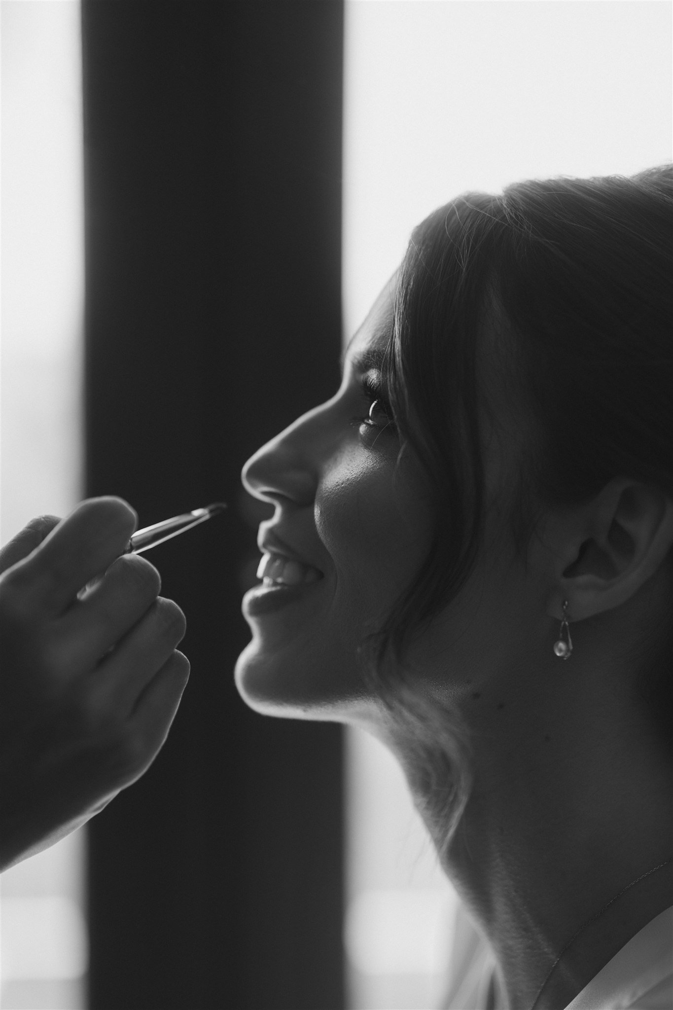 A woman smiling as a person applies makeup with a brush near her face, in black and white.