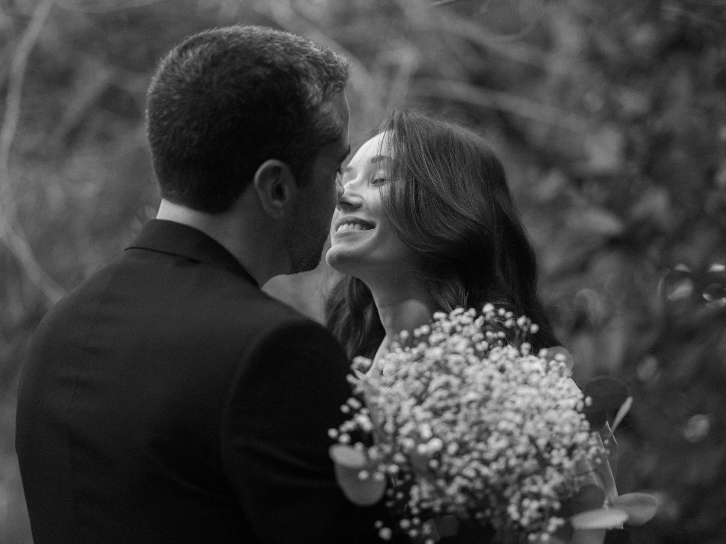 A black and white photo of a couple smiling closely, with the woman holding a bouquet of flowers, likely during a romantic or wedding moment.
