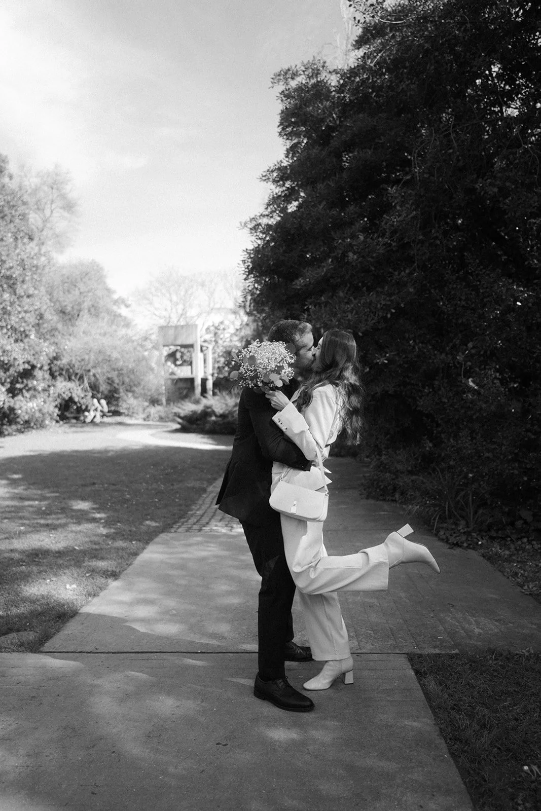 A couple is kissing outdoors, with the woman holding a bouquet of flowers, and she has one leg lifted. It is in black and white, and they are standing on a sidewalk with trees in the background.