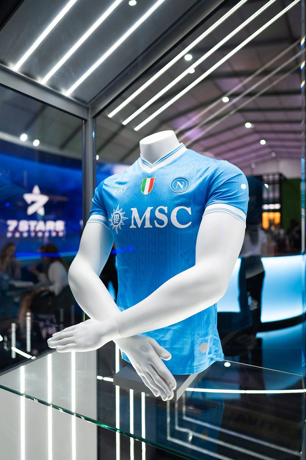 A blue Napoli football club jersey displayed on a mannequin with crossed arms, inside a glass case at an exhibition or store.