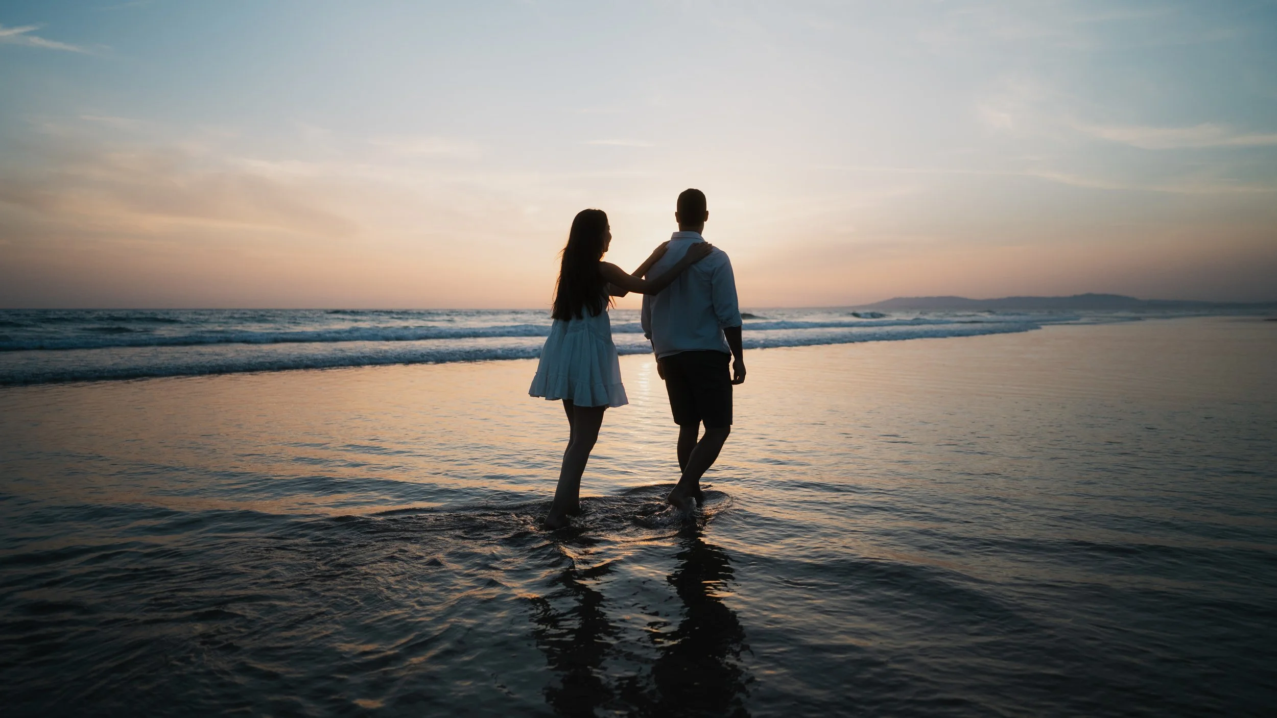 A couple walking hand in hand along the beach at sunset, with the woman touching the man's shoulder, both silhouetted against the colorful sky and calm ocean waves.