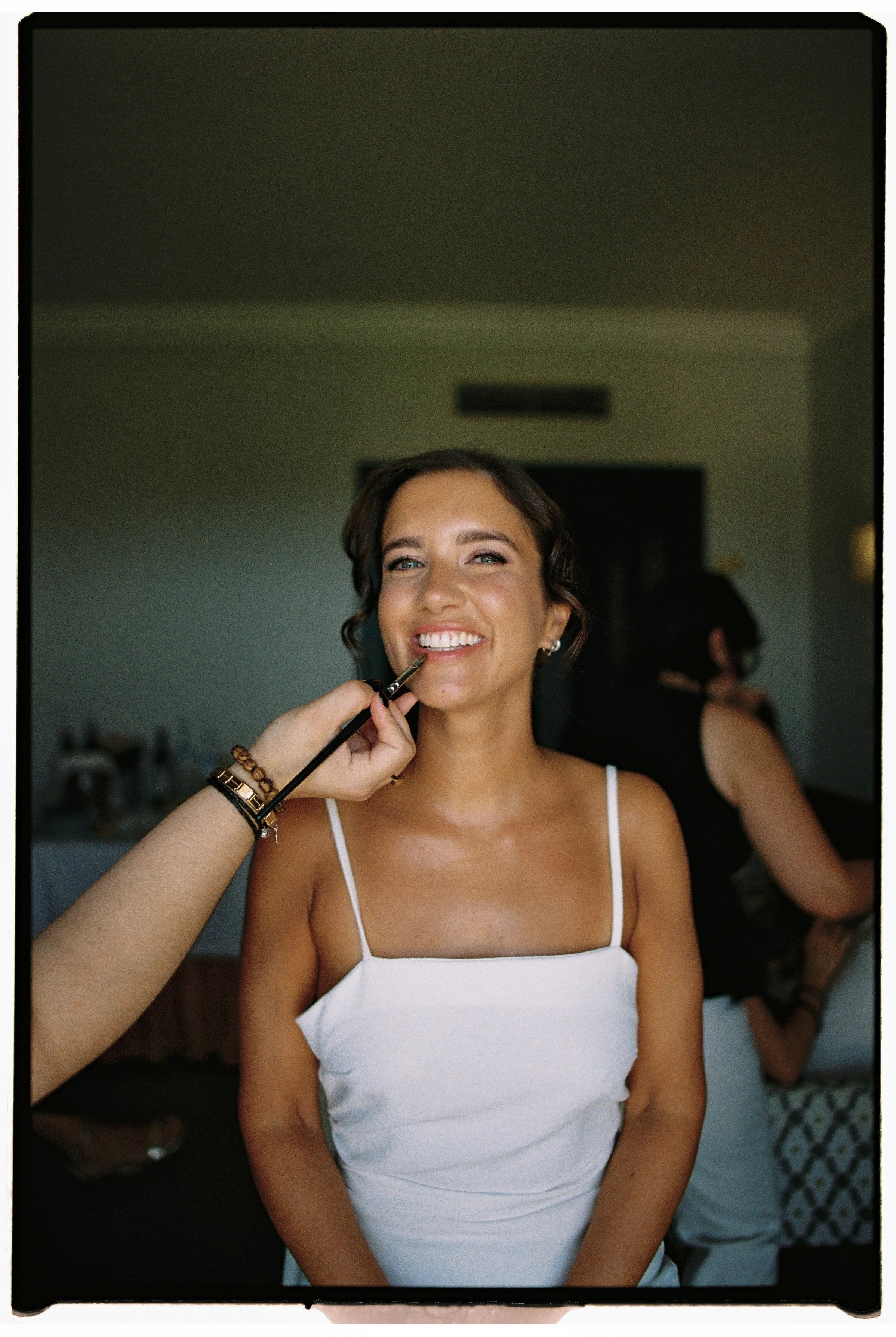 A woman with short hair smiling as makeup artist applies lipstick.