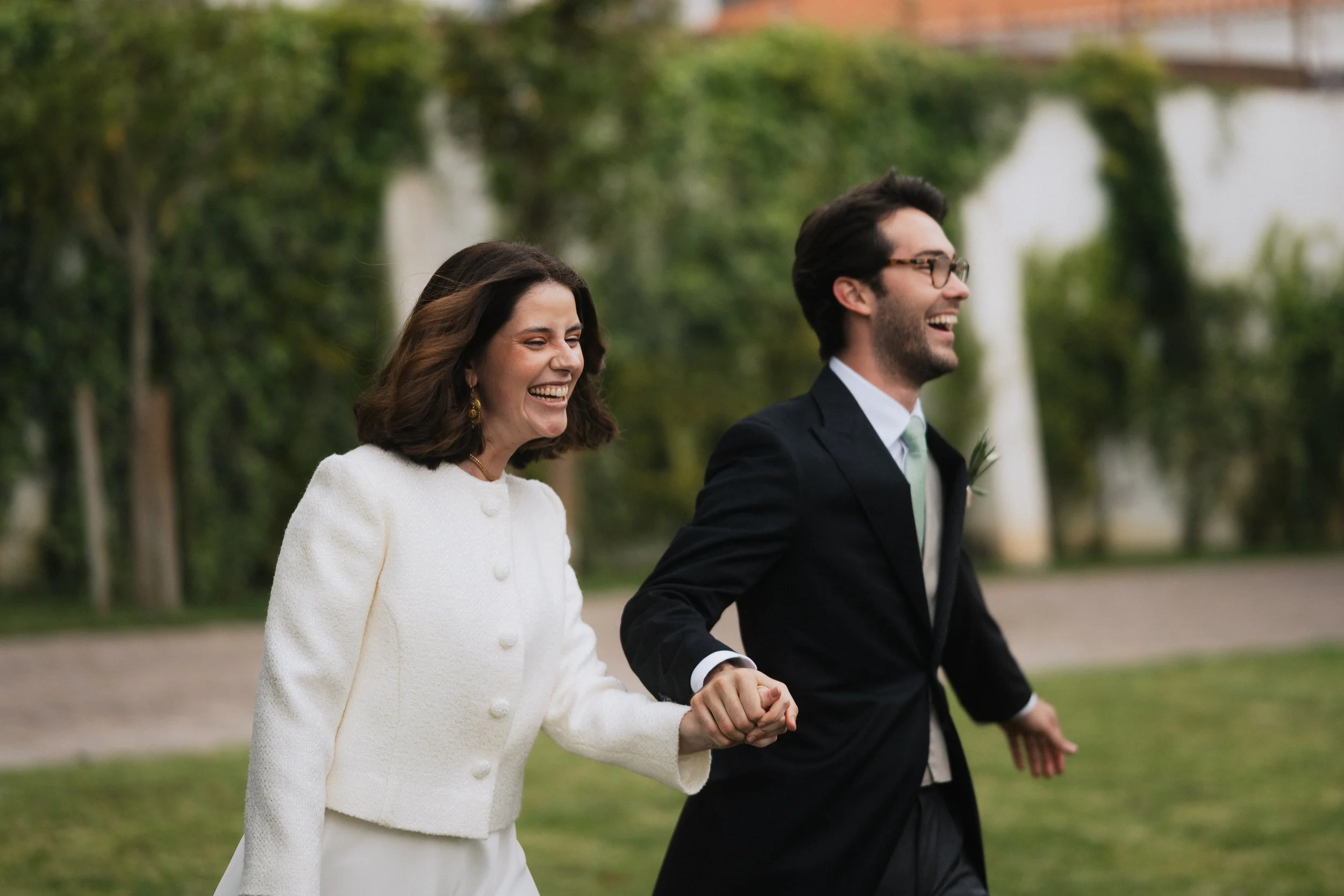 A smiling woman and man dressed in formal attire holding hands while walking outdoors on a cloudy day, with trees and a fence in the background.