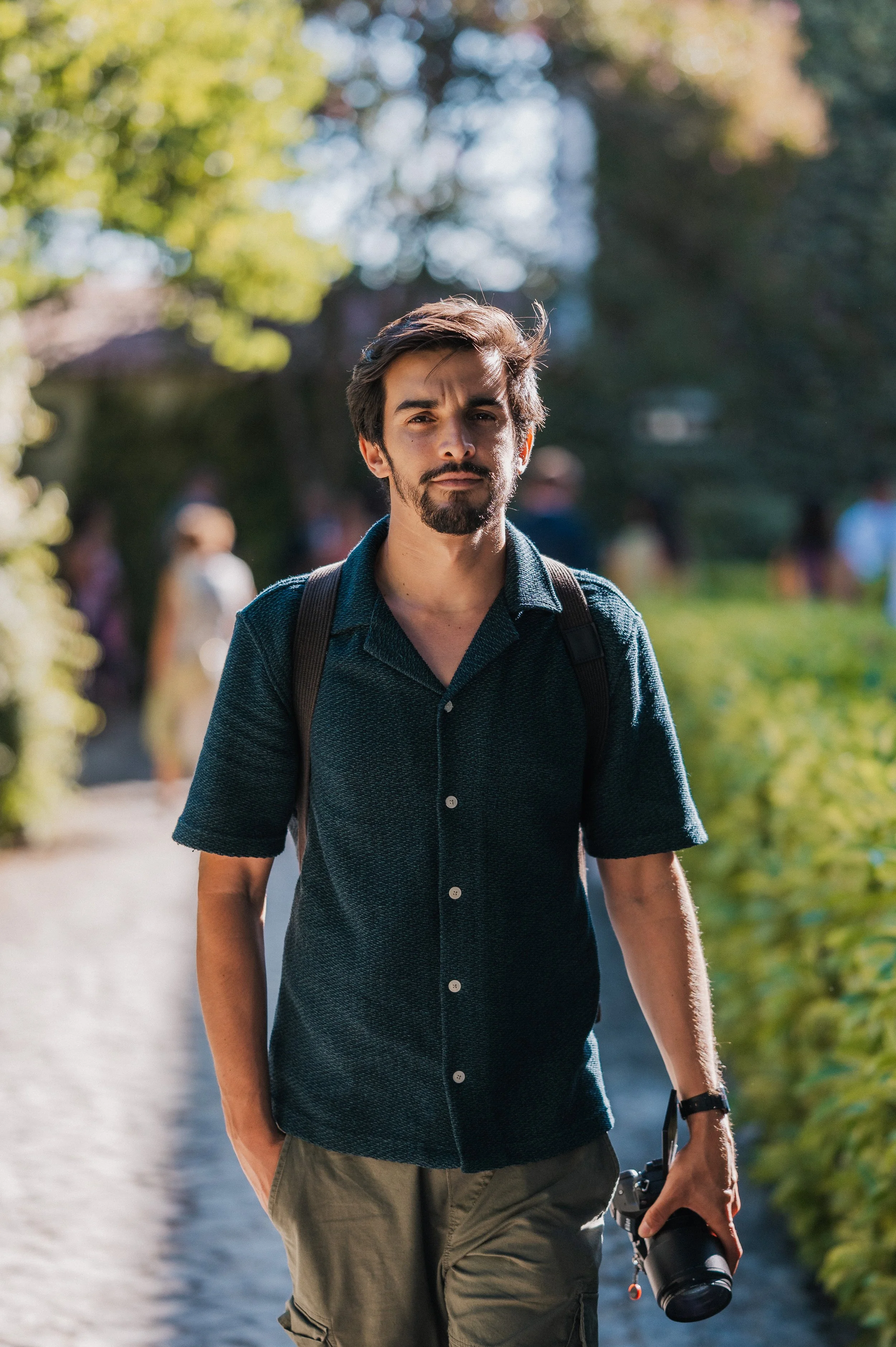 Young man with a camera hanging from his right hand walking outdoors on a sunny day, wearing a dark short-sleeved shirt and a backpack, with blurred people and greenery in the background.