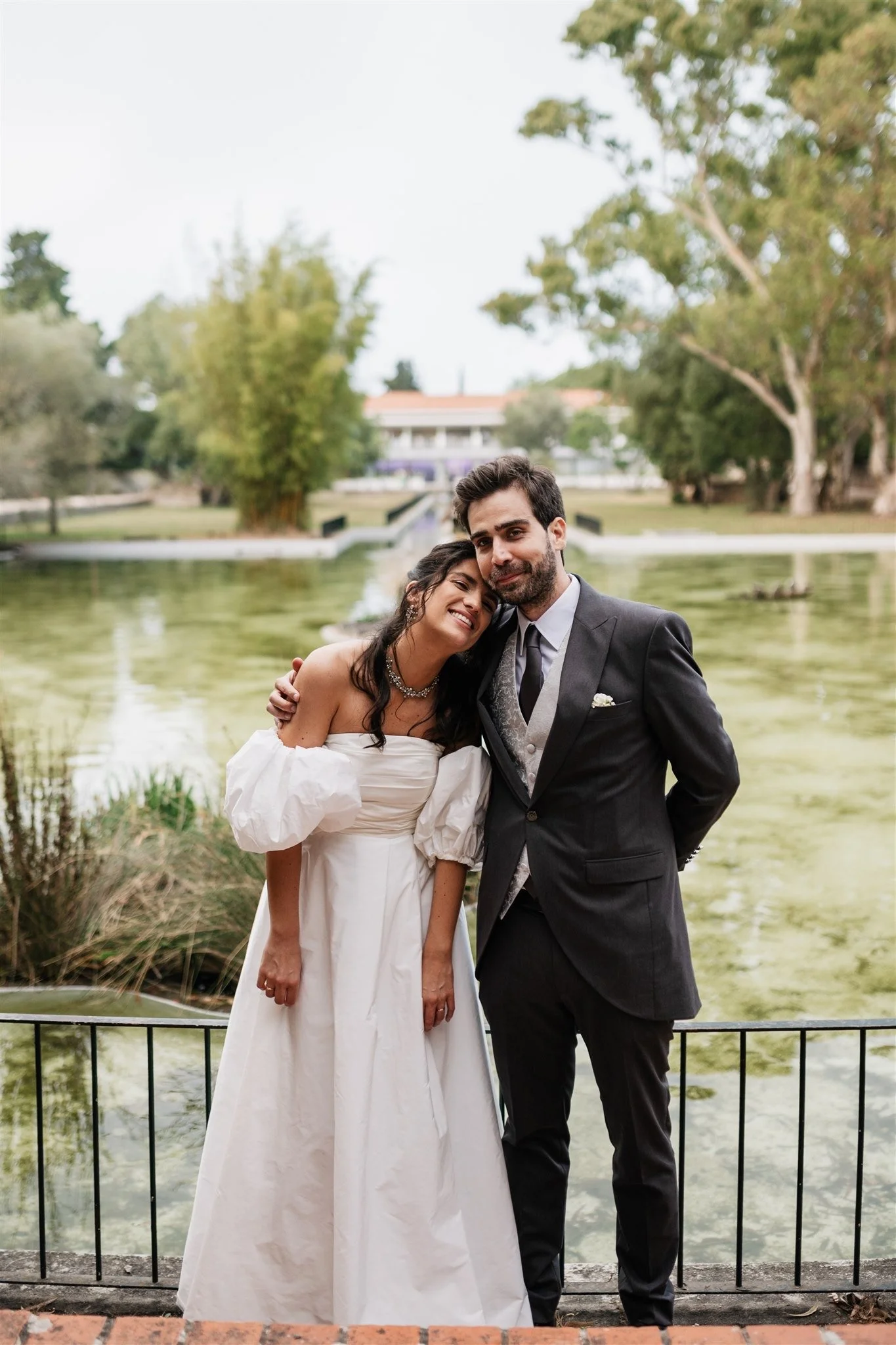 A bride and groom dressed in wedding attire standing by a pond, embracing each other and smiling, with trees and a building in the background.