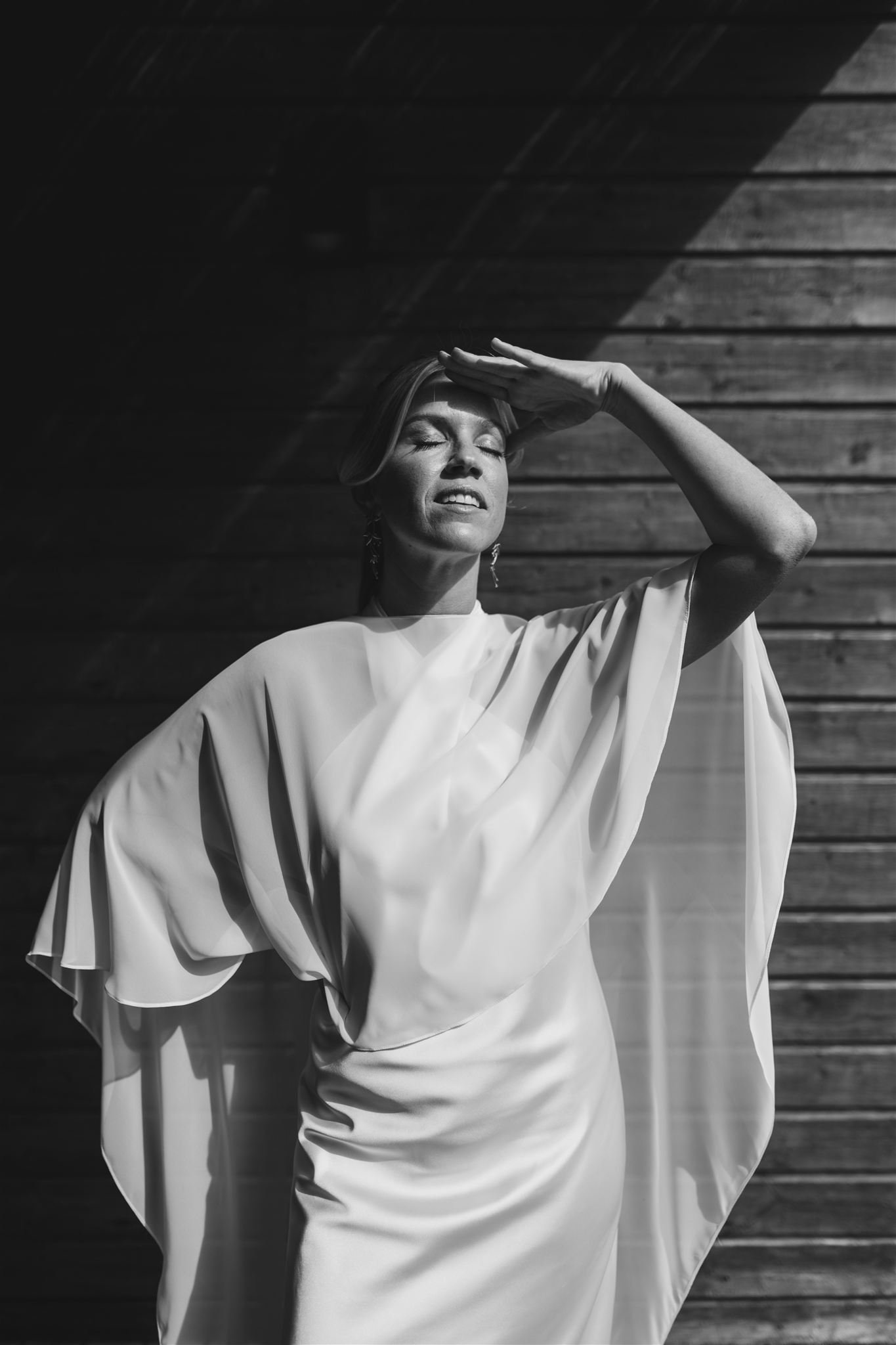 A woman with short hair wearing a loose, light-colored, flowing dress, standing with her eyes closed, one hand on her forehead, against a wooden wall background, in black and white.