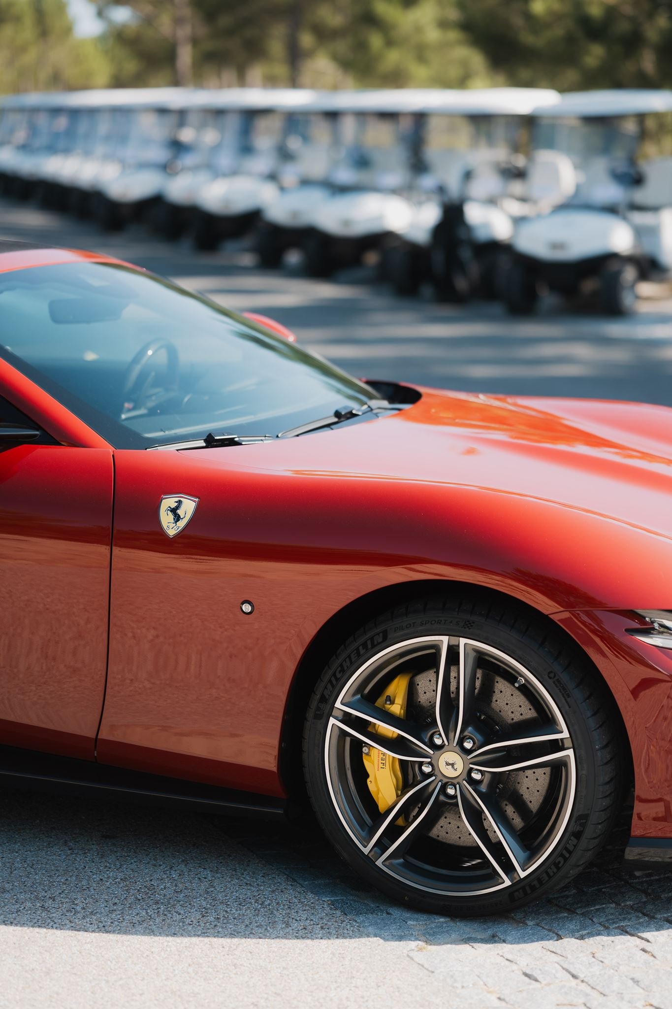 Close-up of the front side of a red Ferrari sports car with a yellow brake caliper, parked on the street, with golf carts and trees in the blurry background.