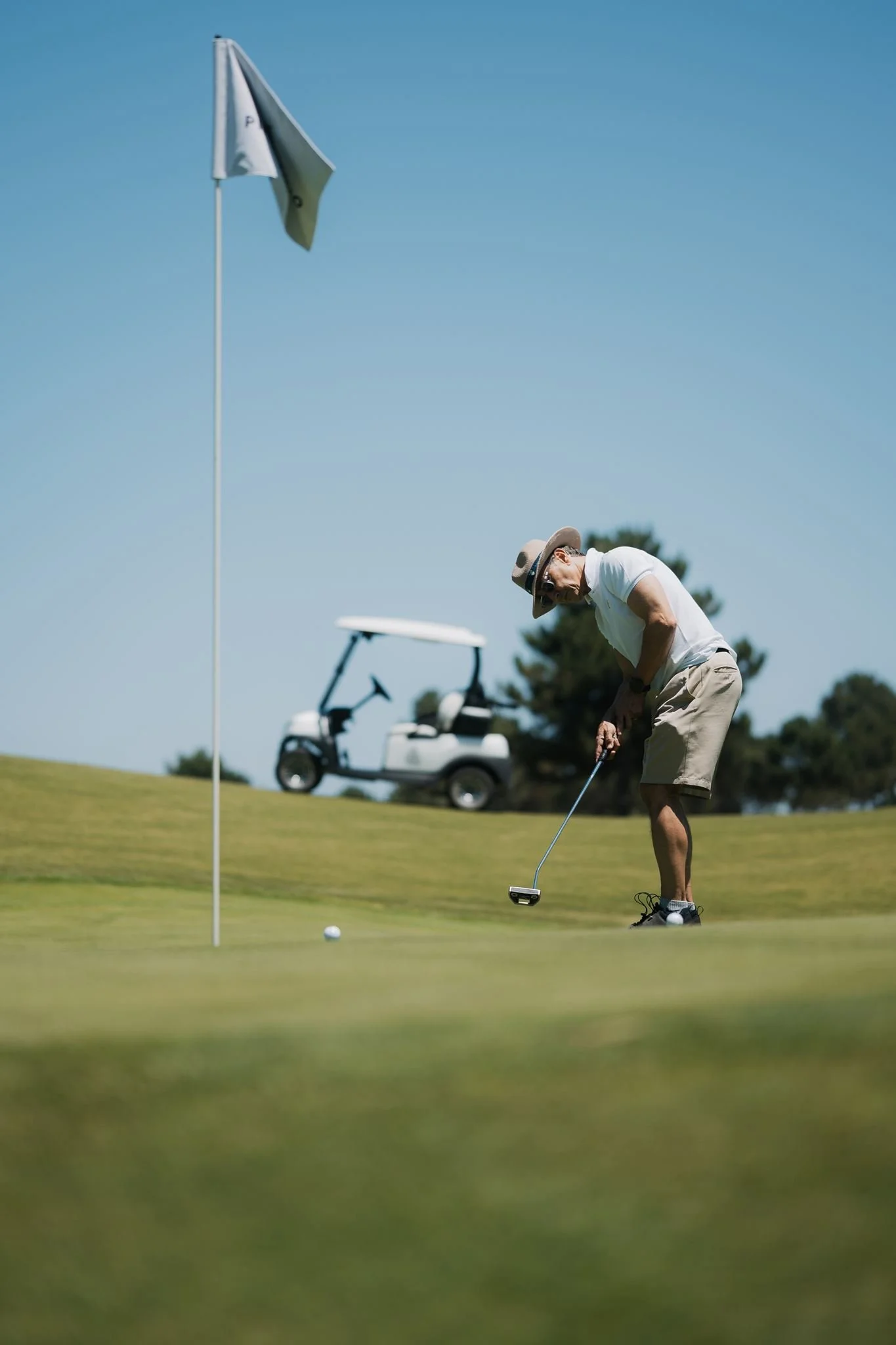 A man wearing a wide-brimmed hat, sunglasses, and a white shirt playing golf on a sunny day. He is bent over, preparing to putt on a green with a golf cart in the background.
