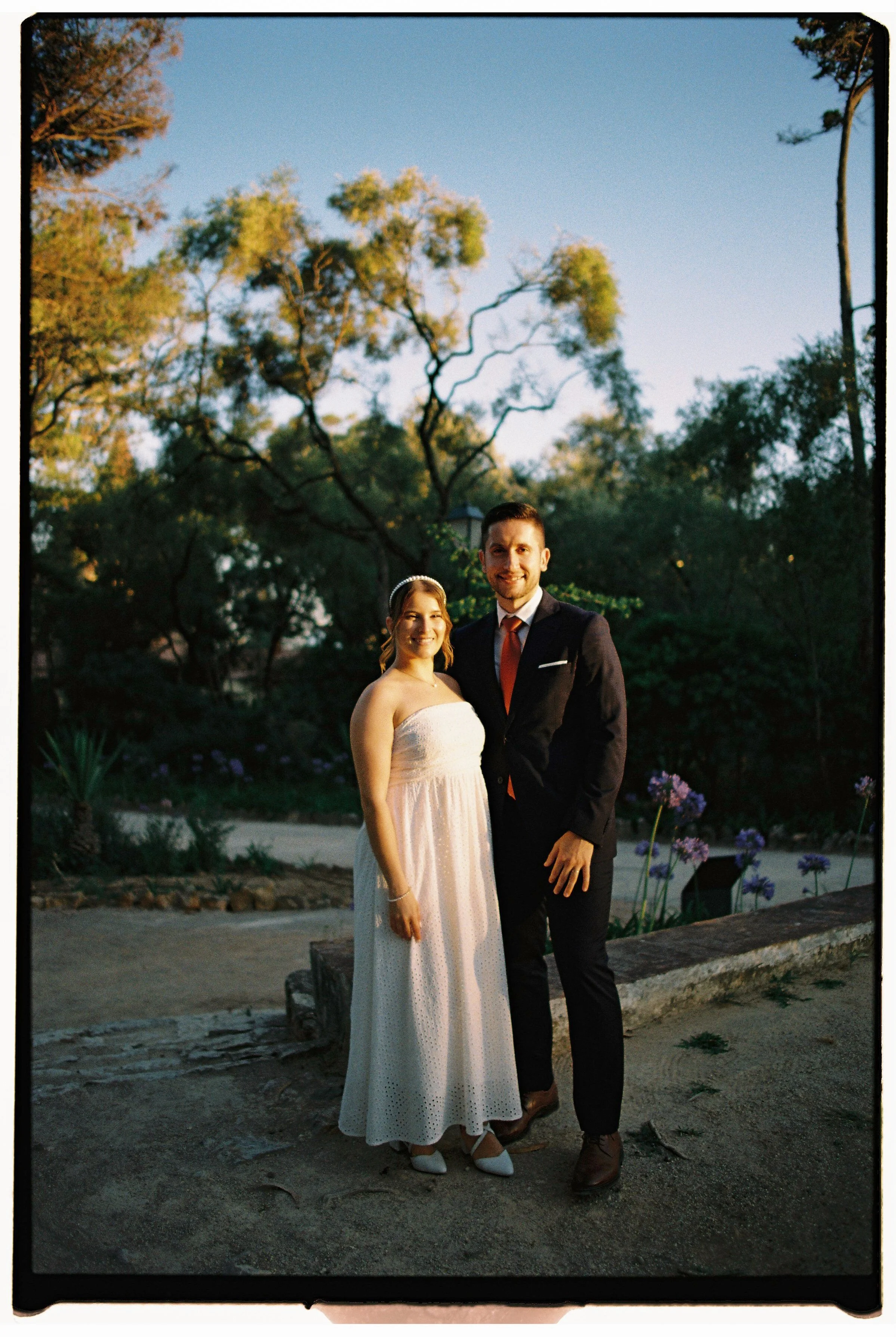 A bride in a strapless white wedding dress and a groom in a dark suit with a red tie, standing outdoors near trees and purple flowers during the evening.