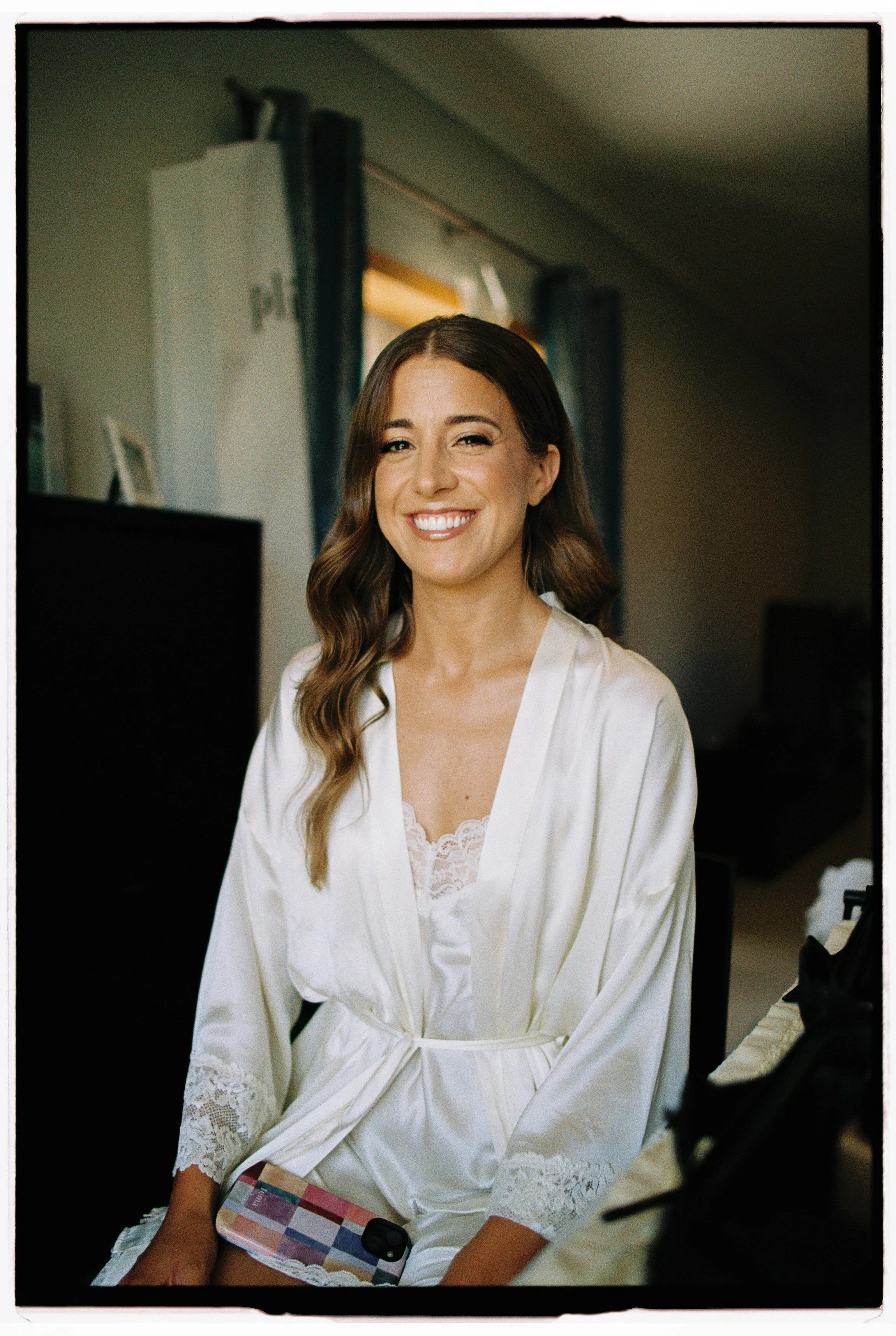 A woman with wavy brown hair smiling and winking while wearing a white satin robe.