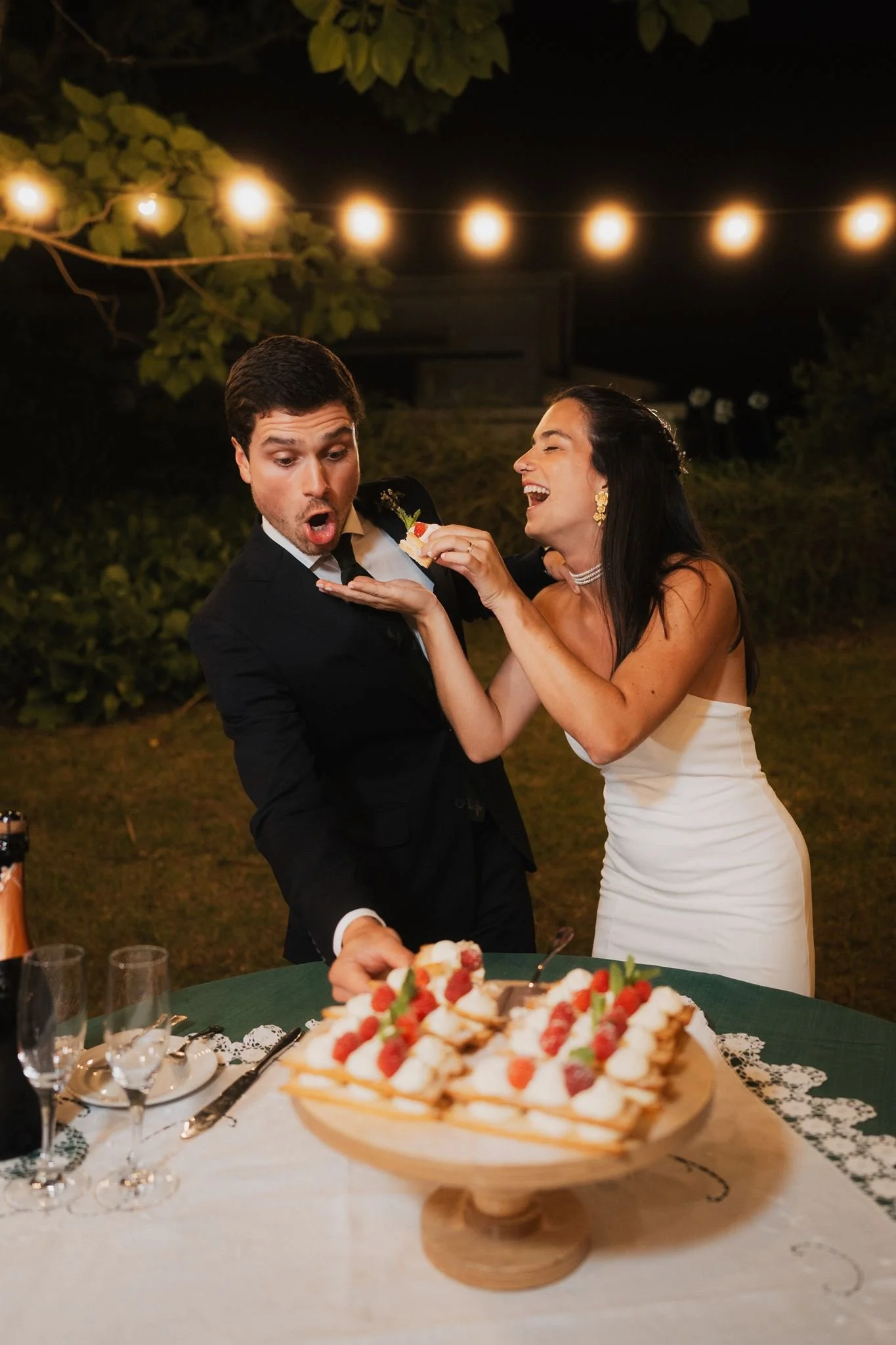 A couple celebrating, with the woman feeding the man a dessert at an outdoor party under string lights. A large cake with strawberries is in the foreground on a decorated table.