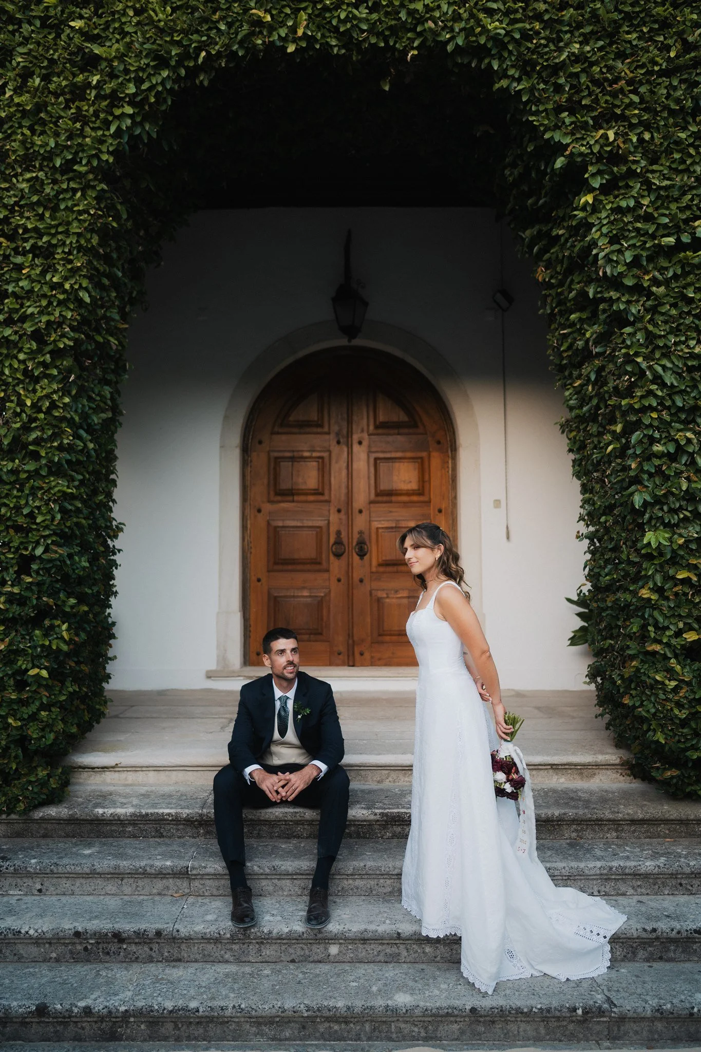 A man in a suit sitting on top of steps and a woman in a white wedding dress standing next to him, outdoors near a wooden door and a green hedge arch, during a wedding.
