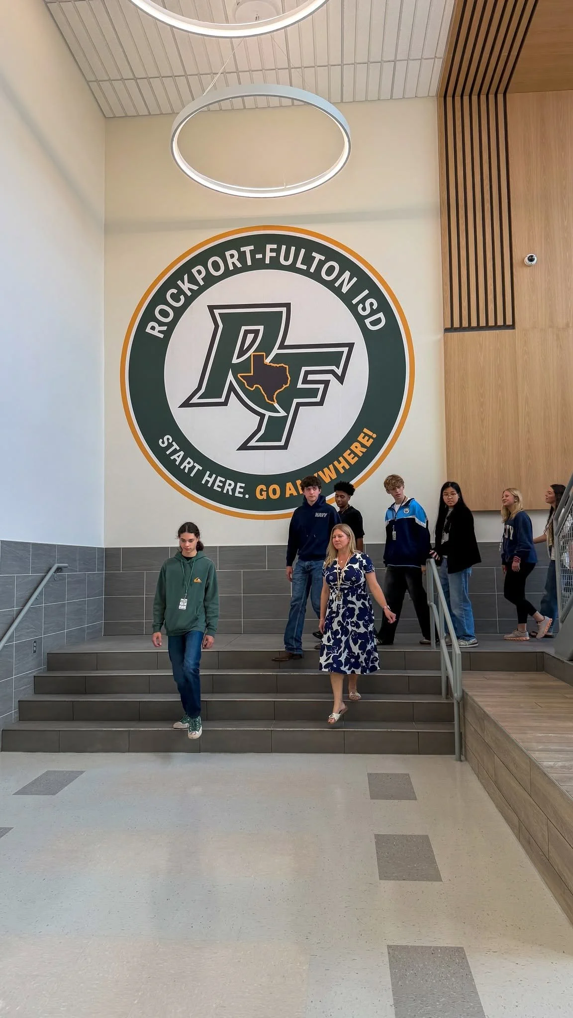 Inside a building with a large logo on the wall that says 'Rockport-Fulton ISD' and 'Start Here. Go Anywhere!' Several people are walking down stairs, including a woman in a blue and white dress and others in casual clothing.