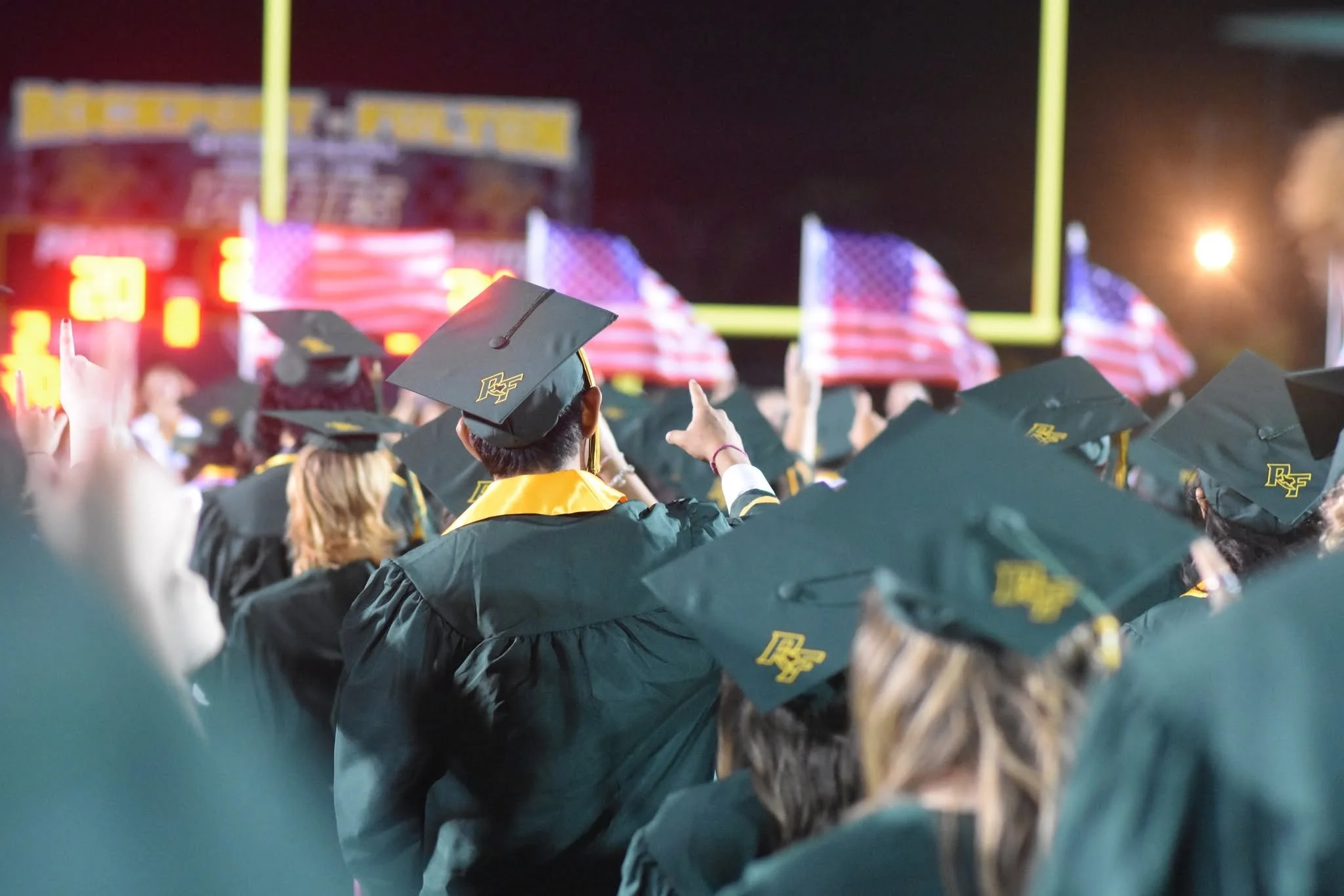 Front row of high school graduates wearing black caps and gowns at a nighttime outdoor graduation ceremony, with American flags in the background and a football field goal post visible.