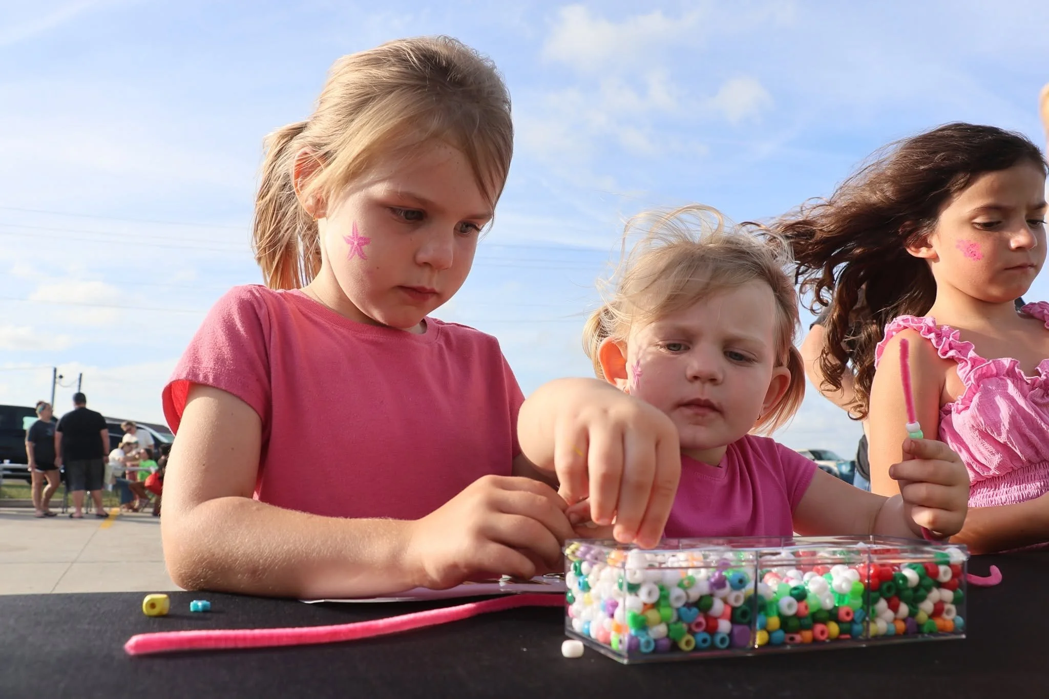 Three young girls with painted faces sitting at a table, playing with beads. The girl on the left has a star painted on her cheek and is wearing a pink shirt. The girl in the middle has a design painted on her cheek and blonde hair. The girl on the right has a flower painted on her cheek and dark curly hair. In the background, there are people and cars outdoors under a partly cloudy sky.