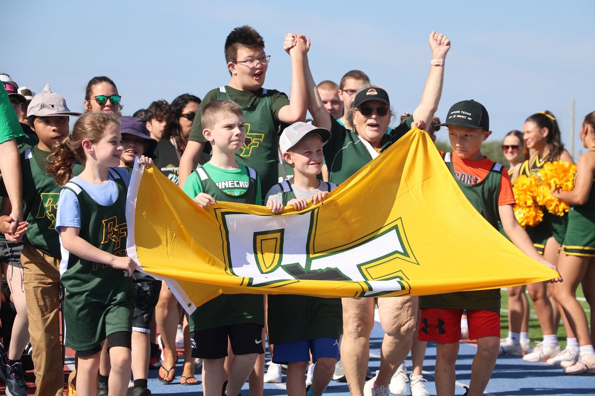 A group of children and adults participating in a sports event outdoors, holding a yellow flag with a logo, with a clear blue sky overhead.