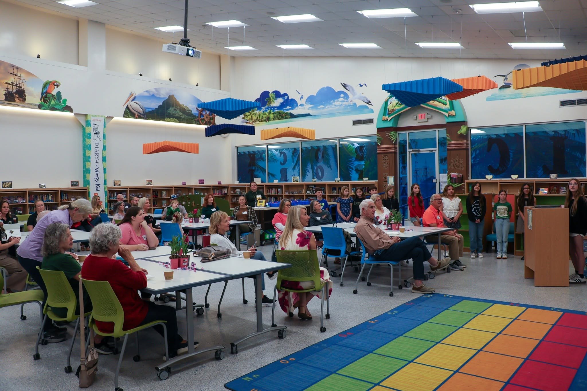 People gathered in a room with colorful ceiling decorations and mural art, seated at tables and standing, watching a presentation or performance near a set of bookshelves.