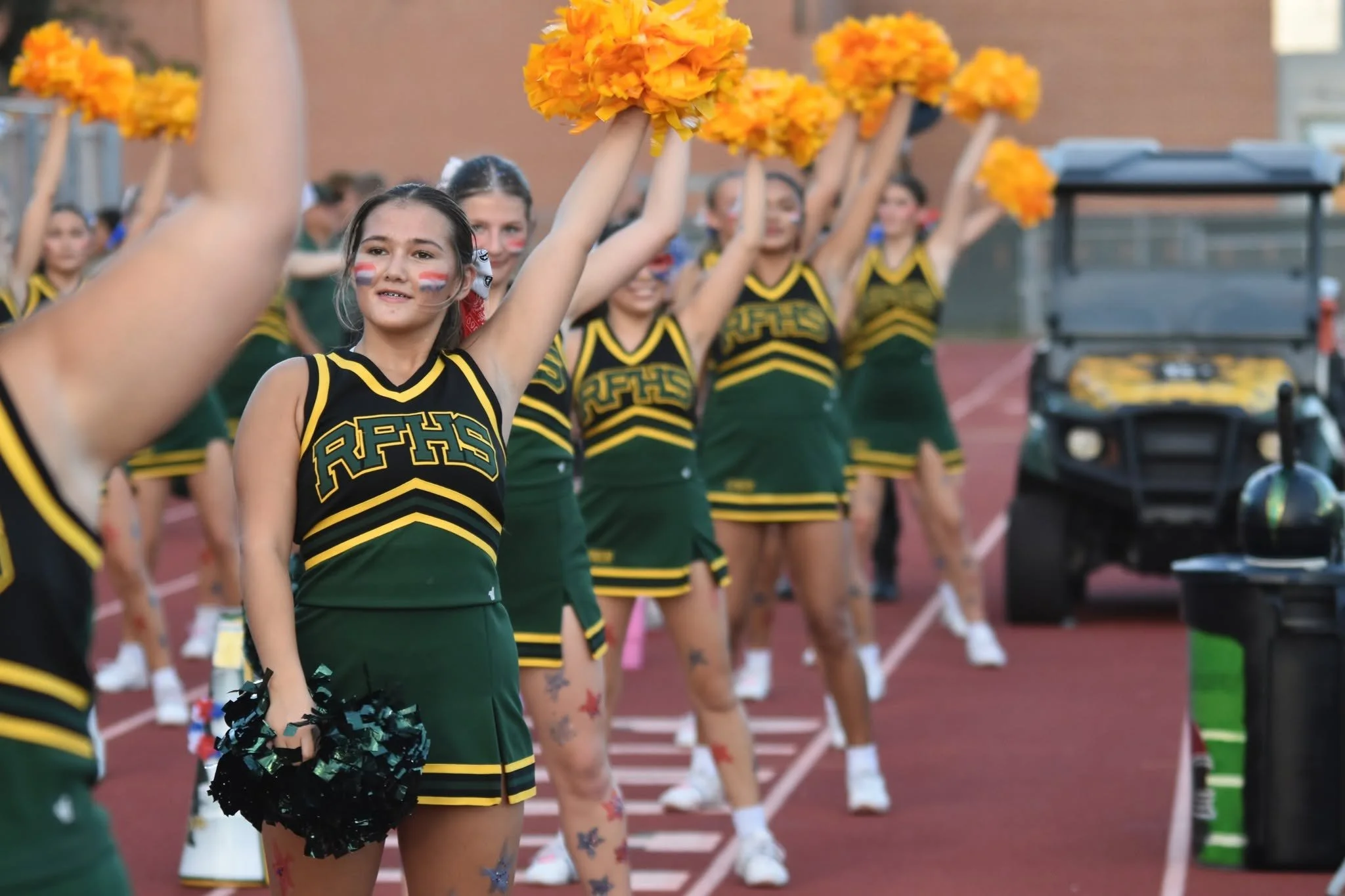 A group of cheerleaders in green and yellow uniforms standing in line on a track, holding black pom-poms, with some wearing face paint and tattoos, raising their arms, possibly at a sports event or parade.