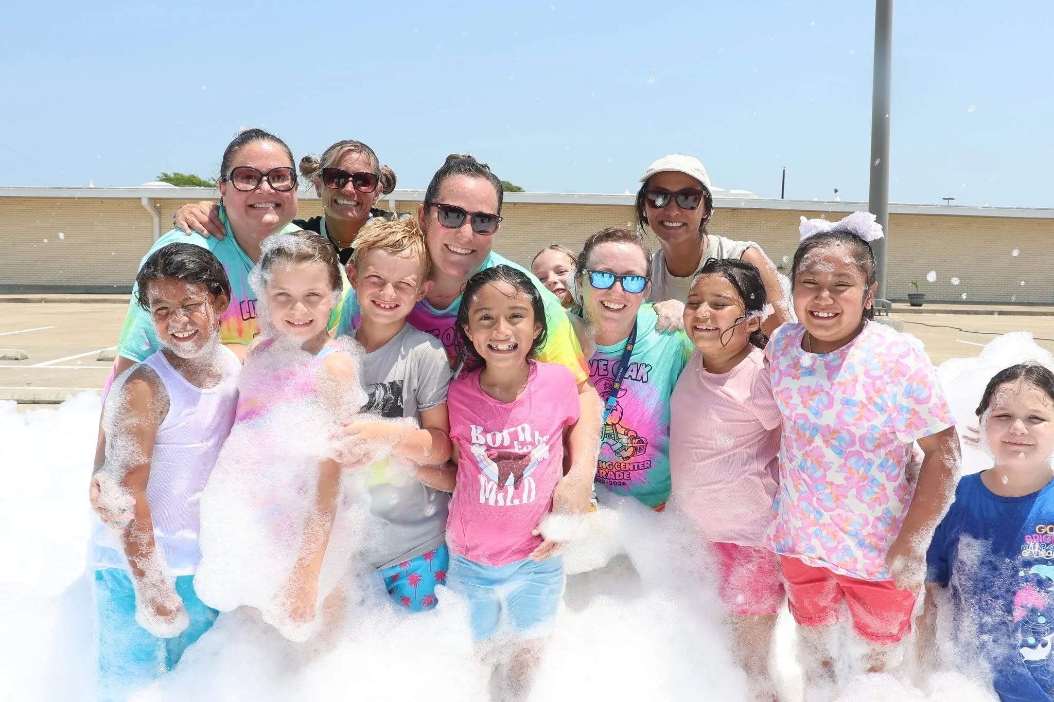 Group of children and adults enjoying a foam party outdoors on a sunny day, smiling and wearing colorful clothes and sunglasses.
